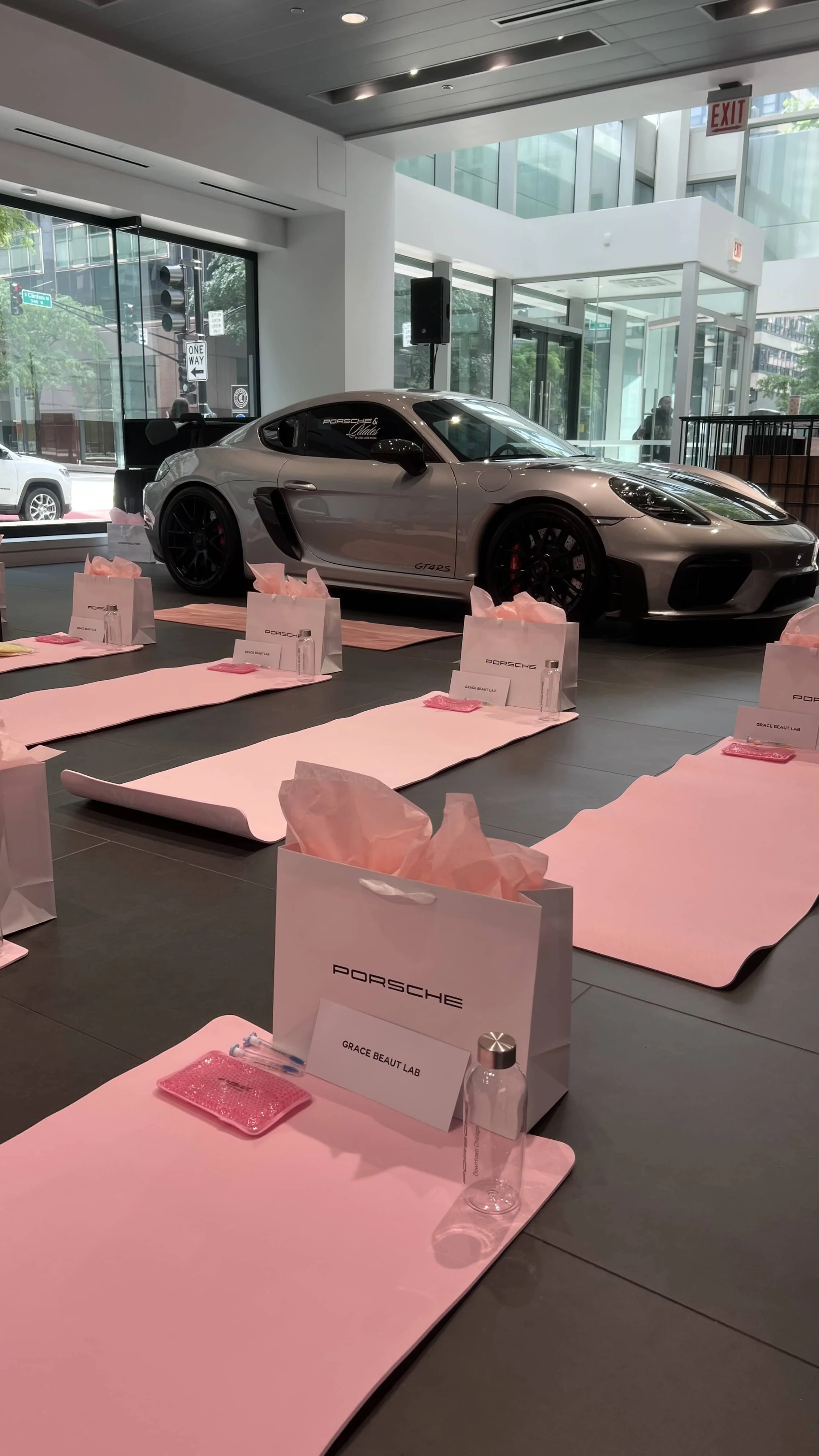 Display of a silver Porsche sports car inside a modern showroom with pink mats, gift bags, and skincare product samples set up on tables in front of the car.