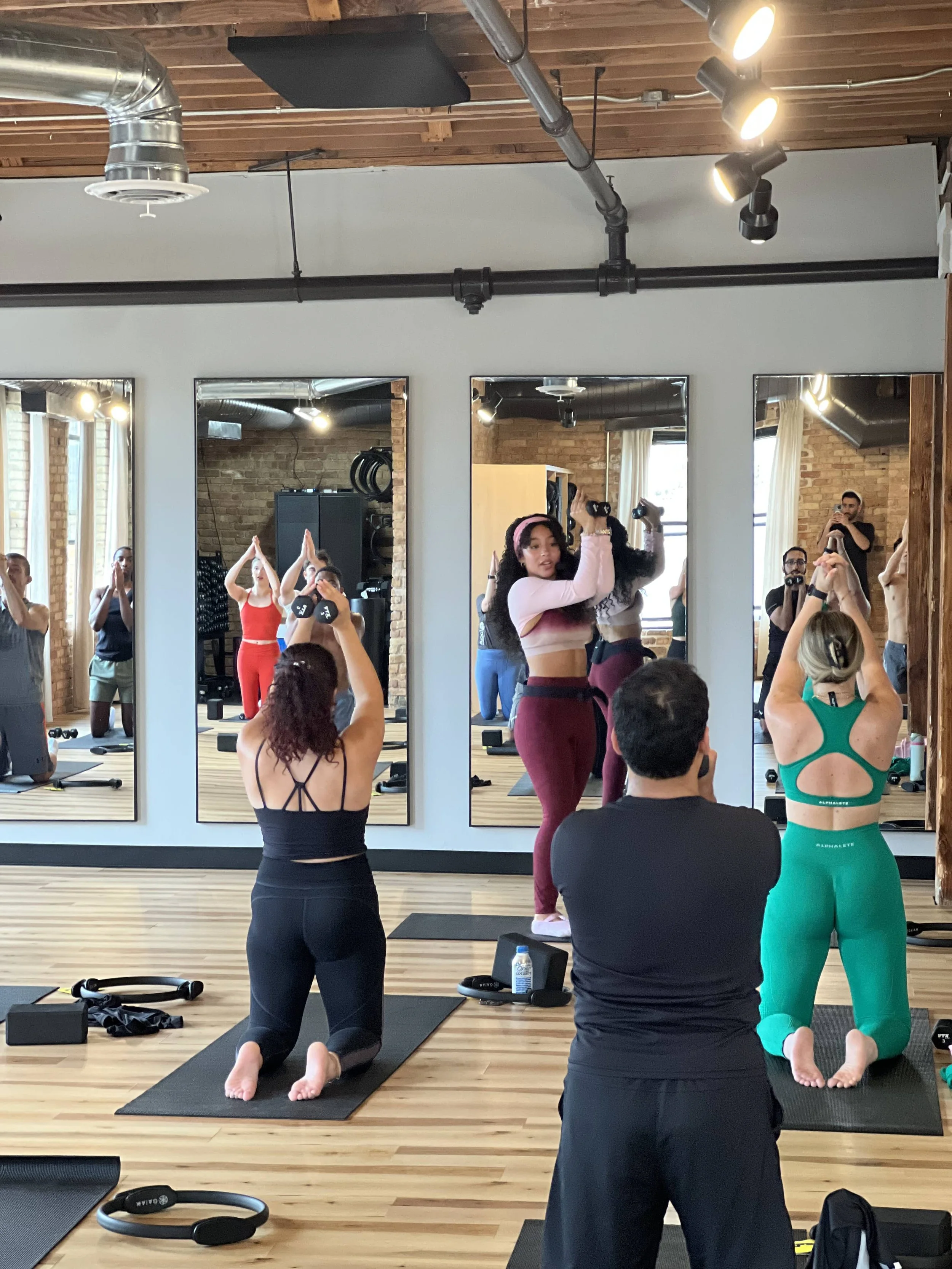 A group exercise class taking place in a fitness studio with wooden floors and exposed brick walls. Participants are doing a kneeling yoga pose on black mats, with some holding dumbbells and others raising their arms or in prayer position. There are 