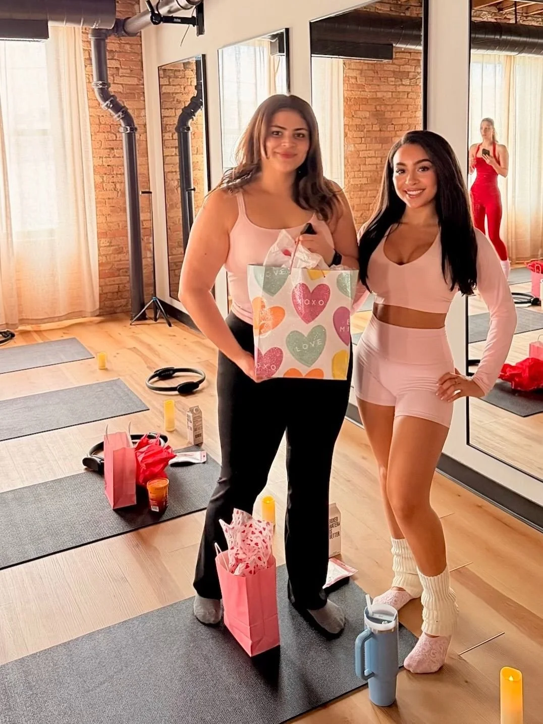 Two young women in workout attire standing indoors, smiling for the camera, with gift bags and exercise mats around them, and a reflection of one woman in the mirror taking a selfie in the background.