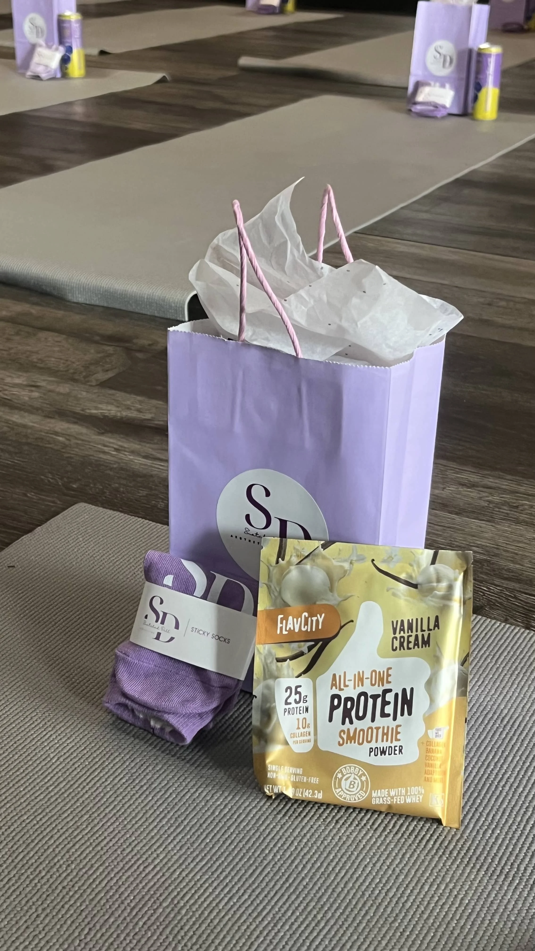 A purple gift bag with pink handles on a table, containing tissue paper. In front of the bag, there is a purple pair of socks, and a packet of vanilla cream protein powder labeled "All-In-One Protein Smoothie" with 25g of protein, 10g of collagen, an