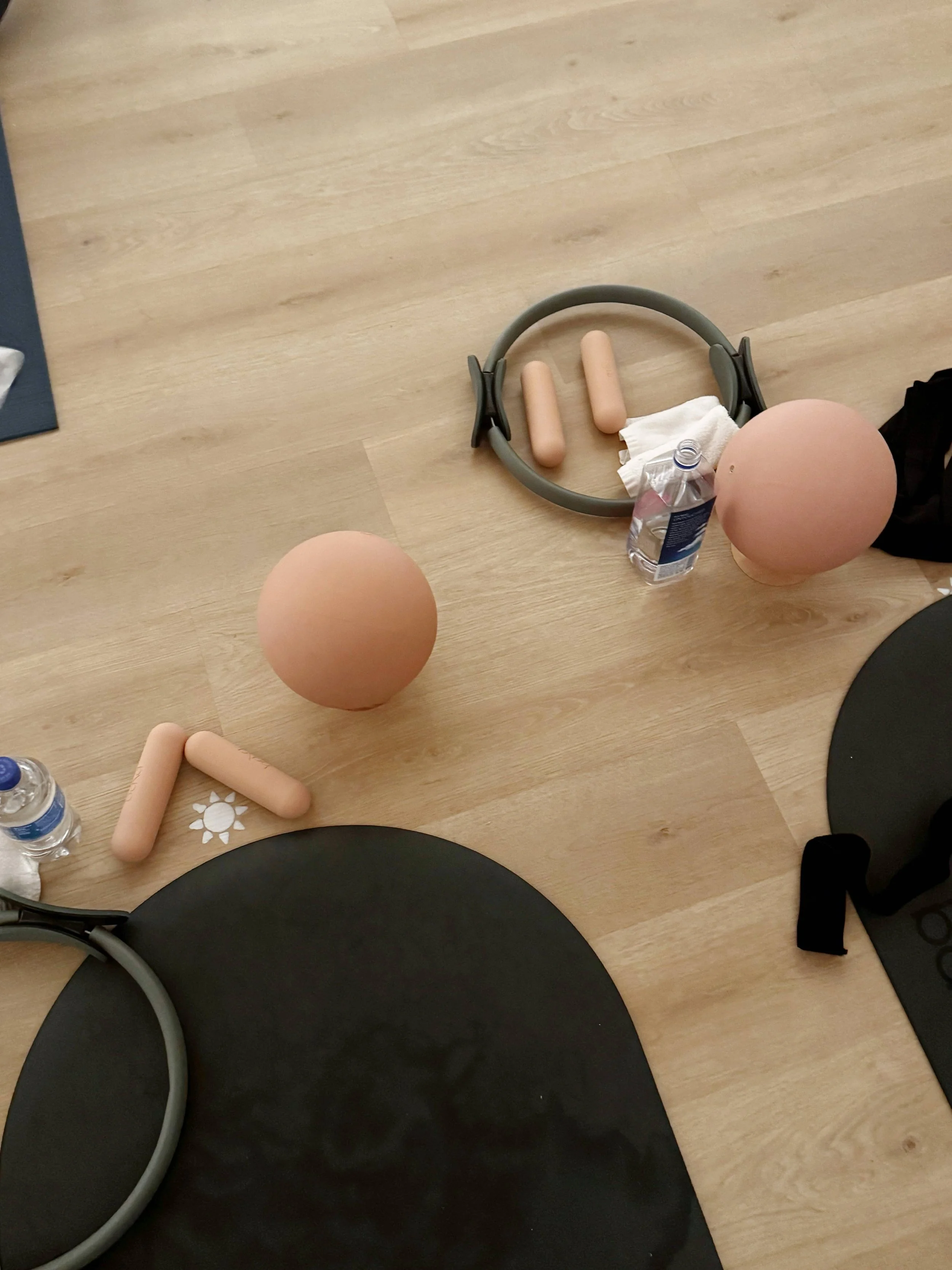 A wooden table with Pilates exercise equipment, including a gray headband, two foam rollers, a water bottle, a towel, and black placemats with black chairs.