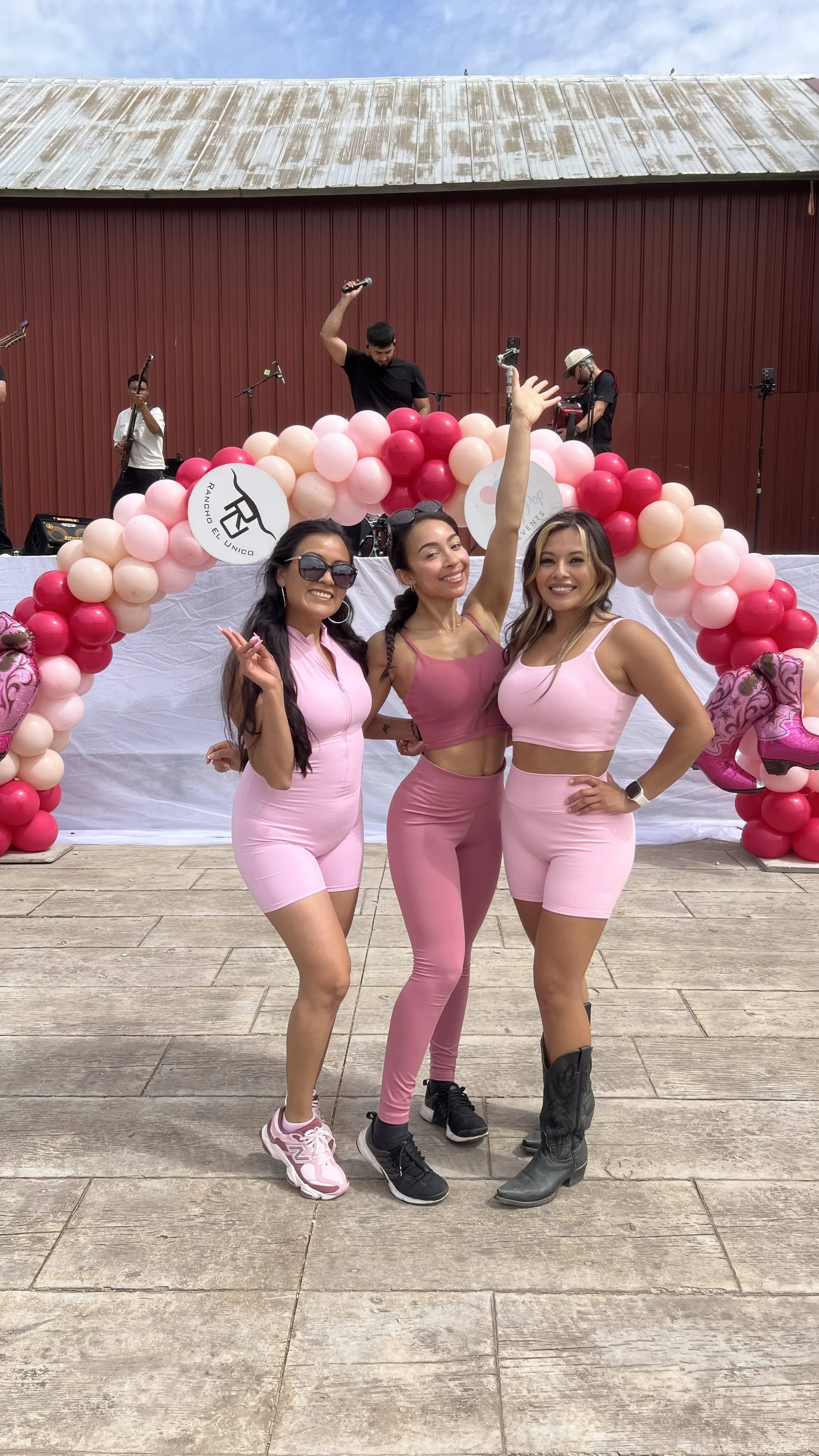 Three women in pink outfits posing at a celebration with a balloon arch and a band performing on stage behind them.