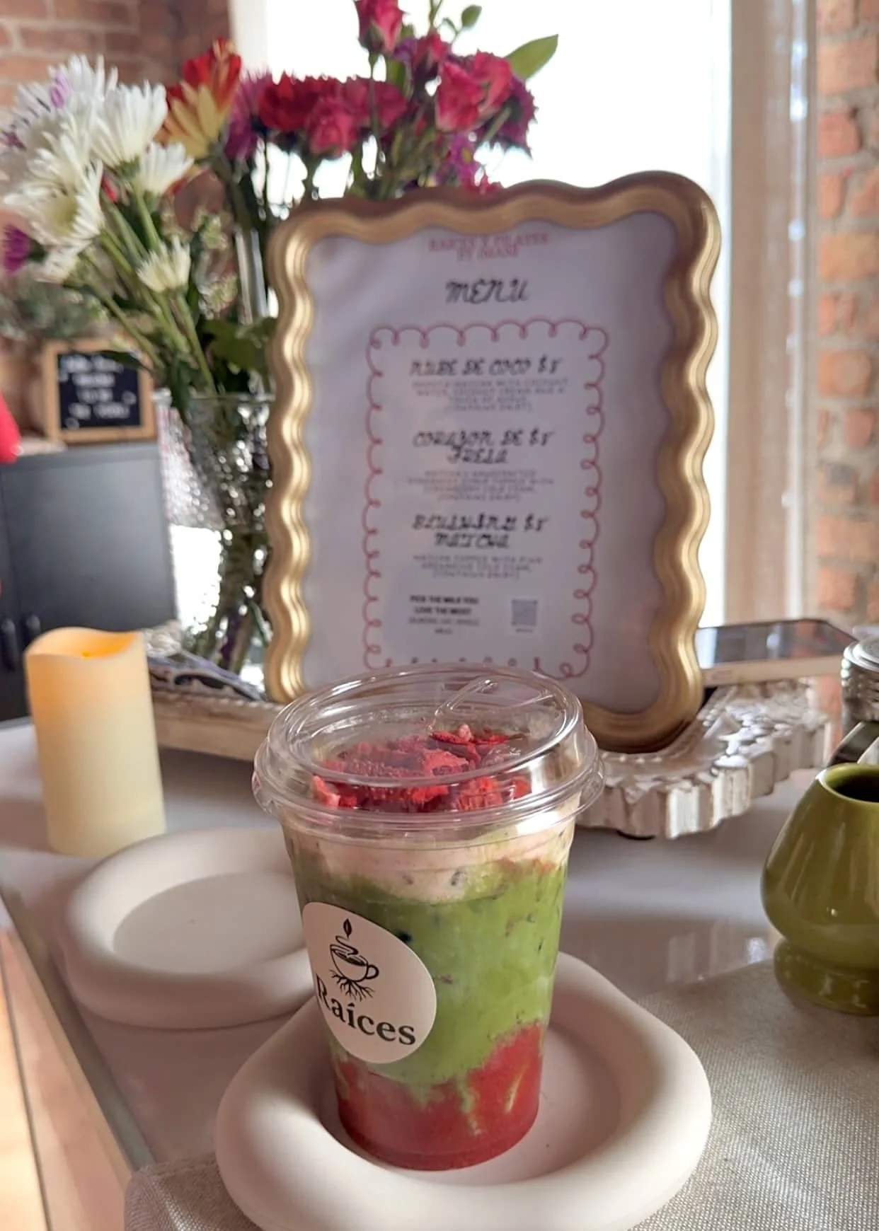 A layered smoothie with green, red, and yellow colors in a clear plastic cup with a lid and stir stick, placed on a white tray with a small white bowl underneath. In the background, there is a floral arrangement with pink, white, and purple flowers, 