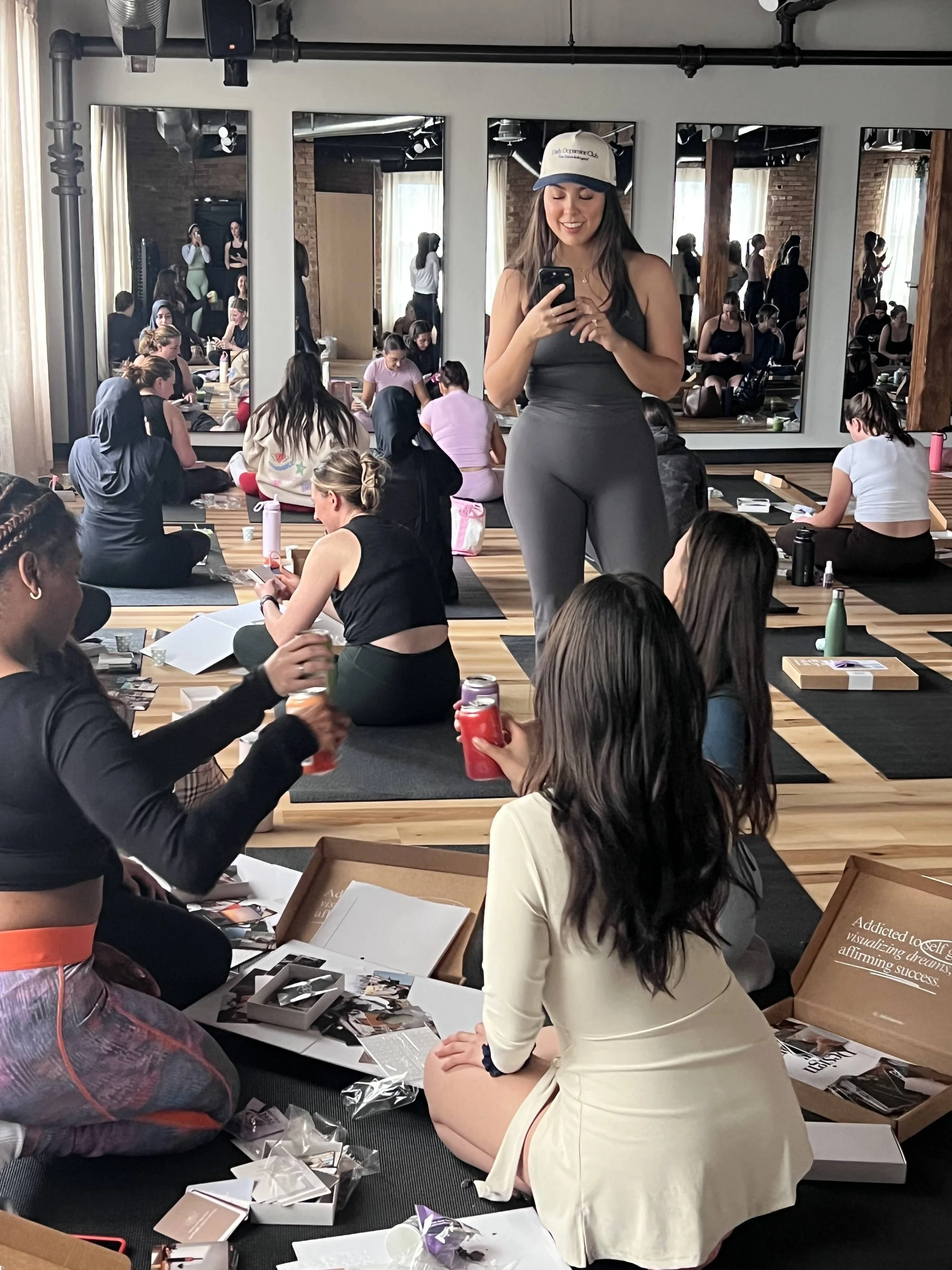 A group of people sitting on the floor in a yoga or meditation class, with a woman in the foreground smiling and looking at her phone, surrounded by open boxes and scattered photographs or cards.
