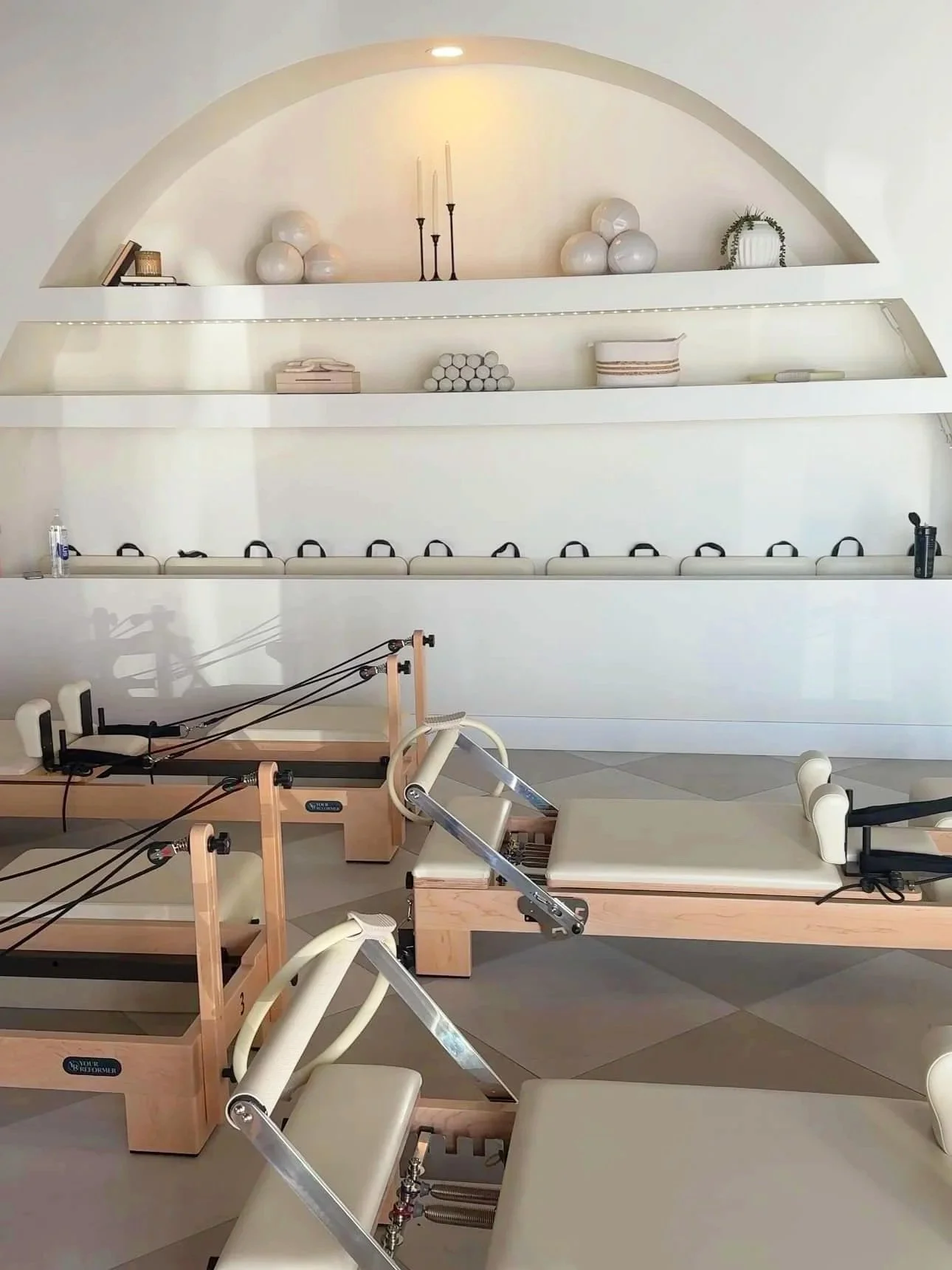 Gym or physical therapy room with Pilates reformer machines and wall shelves decorated with white vases, candles, books, and plants.