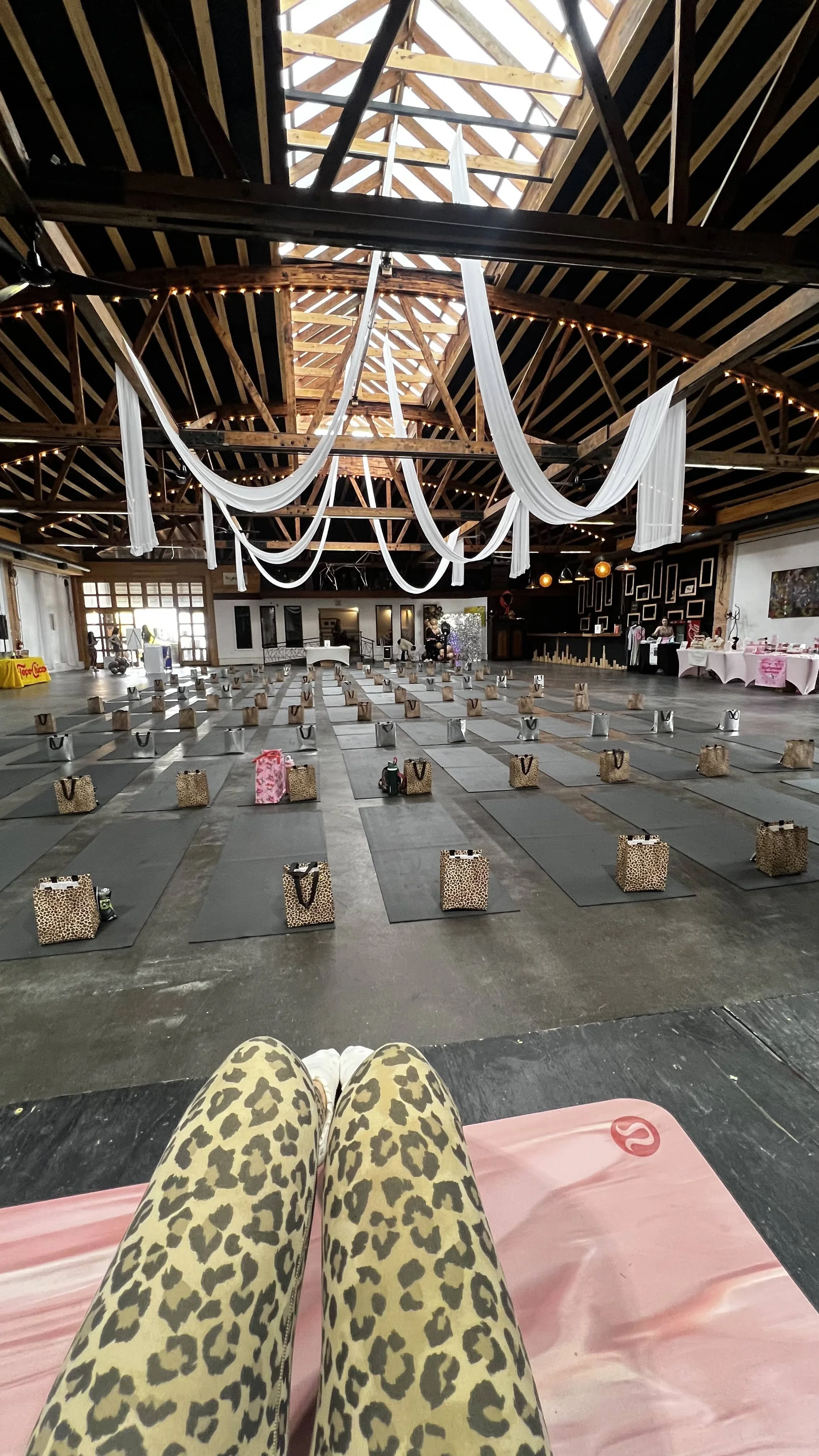 Interior of a large hall decorated for a yoga or fitness class, with mats arranged in rows, gift bags placed on each mat, draped fabric hanging from the ceiling, and exercise equipment along the walls.