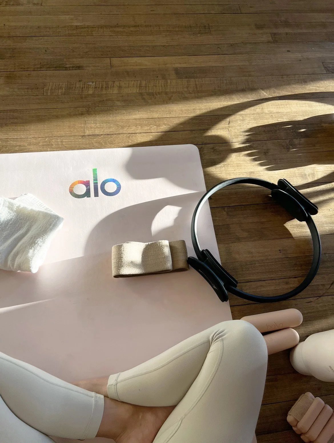 Yoga mat with alo logo, headphones, yoga blocks, and a person's arm in yoga pose on wooden floor.