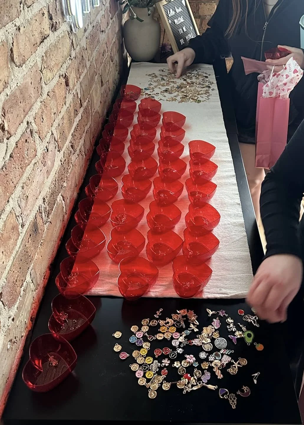 A table with numerous small red heart-shaped bowls, colorful decorative pins, and jewelry pieces on a white cloth, set against a brick wall background.