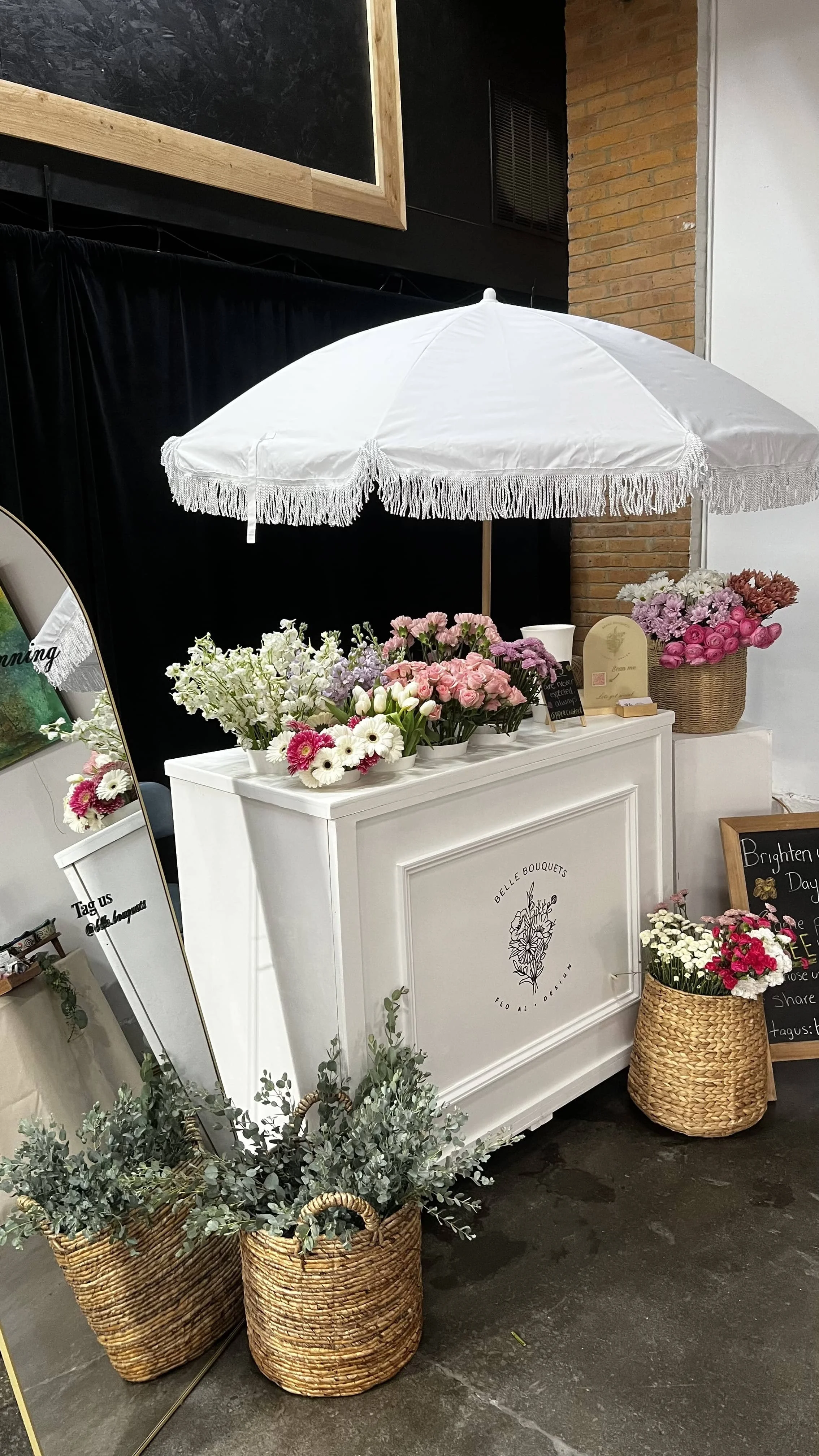 Flower shop display with various pink and white flowers in wicker baskets, white counter, and a white parasol with fringe, in a decorated interior space.