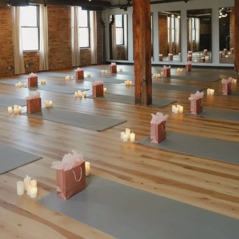 Yoga mats with pink gift bags and candles arranged on wooden floor, large windows with curtains, brick walls, mirrored wall, and wooden beams in the background.