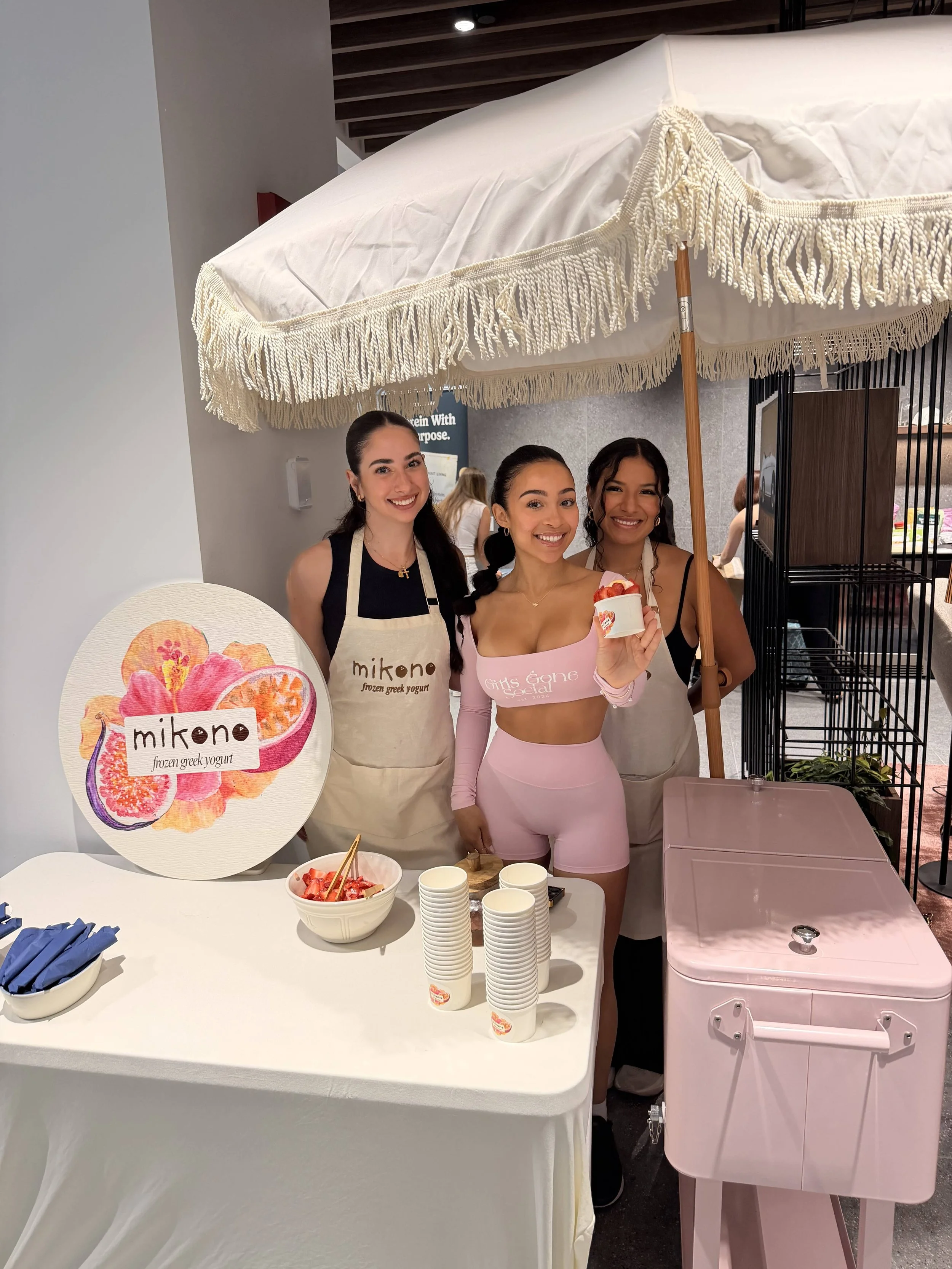 Three women smiling at a frozen yogurt stand, one holding a cup of frozen yogurt, with a sign displaying the Mikone brand logo and illustrations of fruit, and a table with bowls of strawberries and cups of yogurt in front of them.
