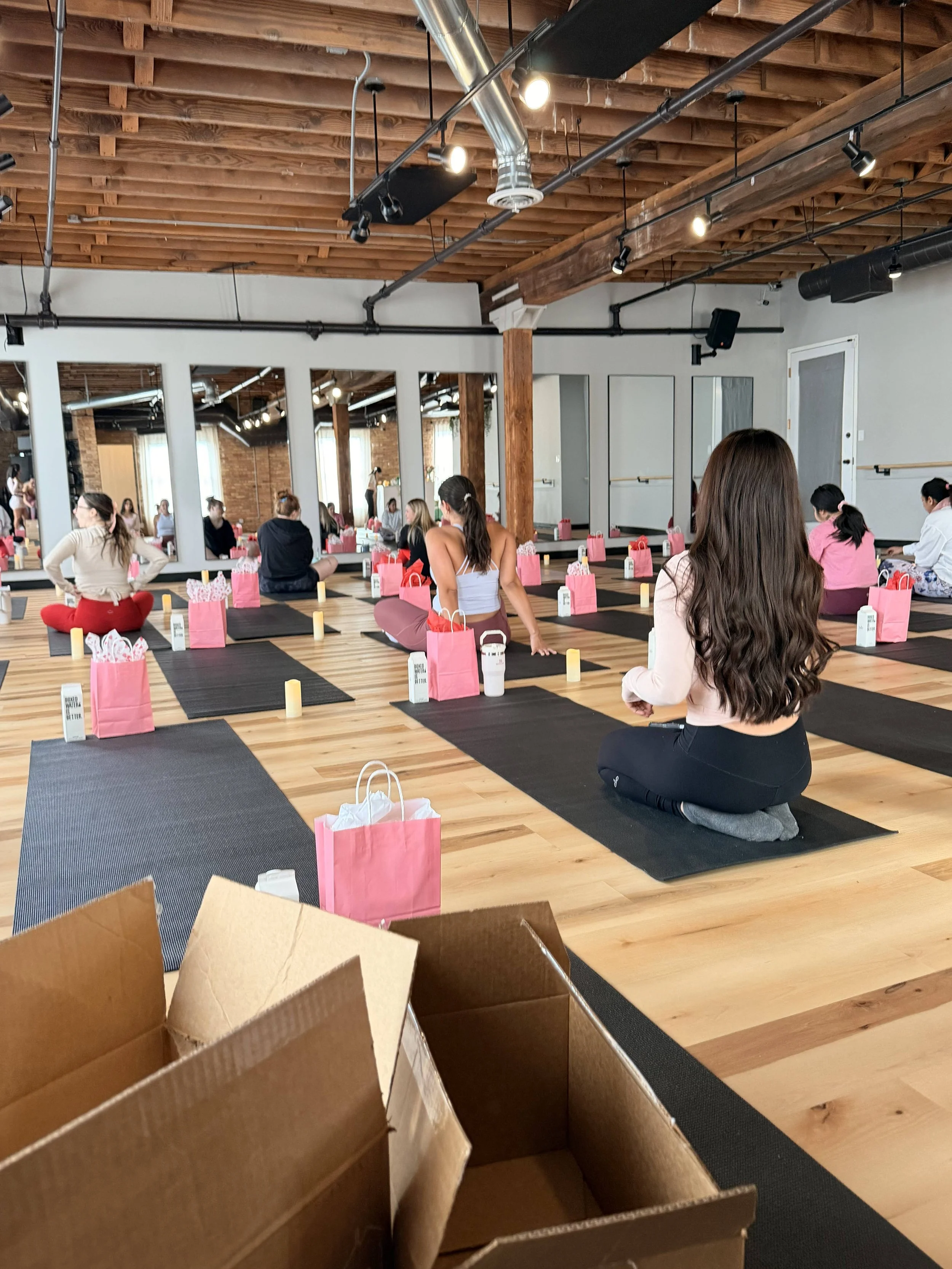 A group of people practicing yoga in a spacious, indoor studio with wooden floors, exposed ceiling beams, and a row of mirrors along one wall. They are seated on yoga mats, with pink gift bags in front of them, and are facing away from the mirrors.