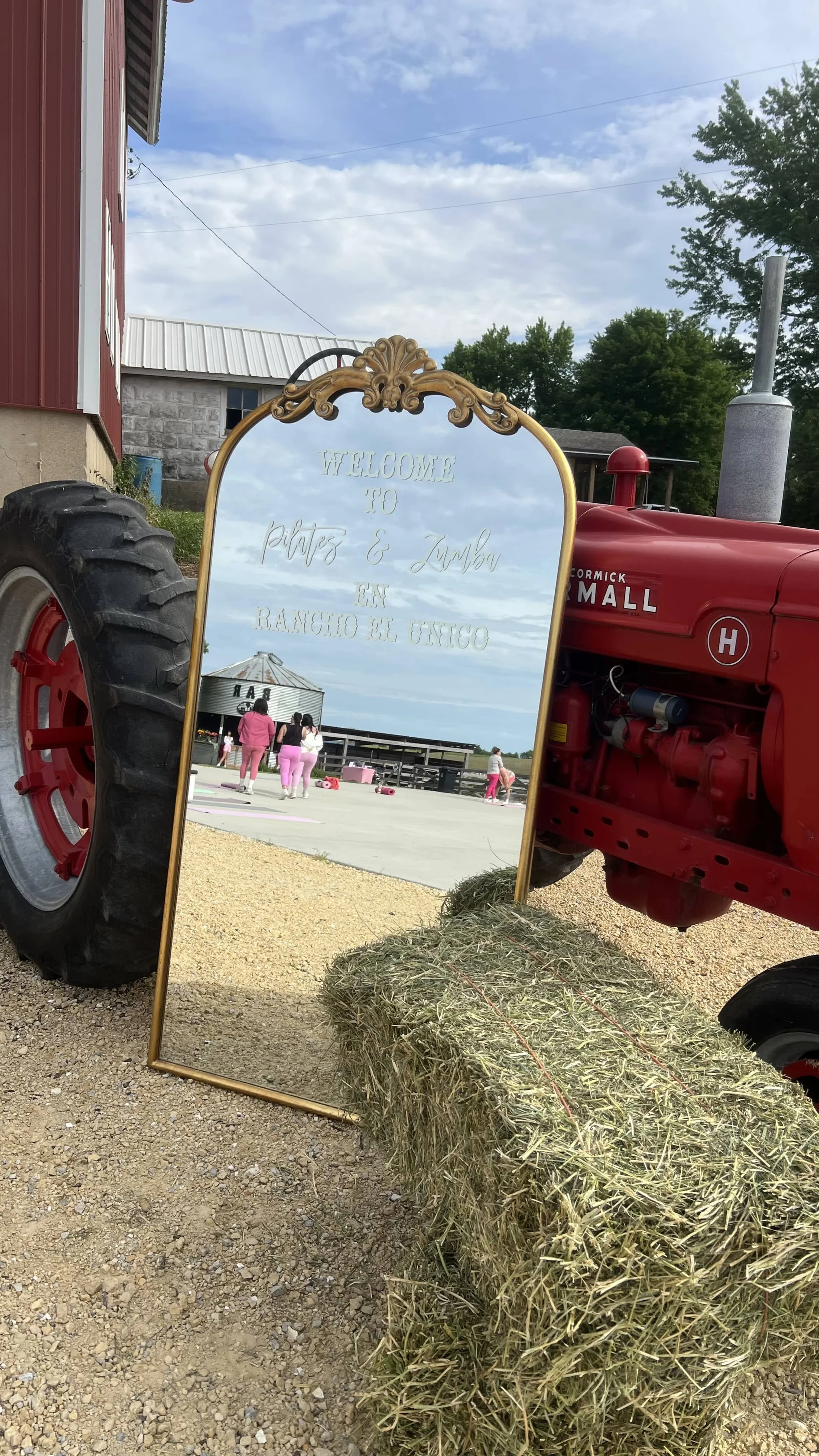 A vintage red tractor positioned next to a decorative mirror that reflects a group of people in pink outfits standing near a barn. There are hay bales on the ground in front of the tractor, and the scene is set on a farm with clear weather and some c