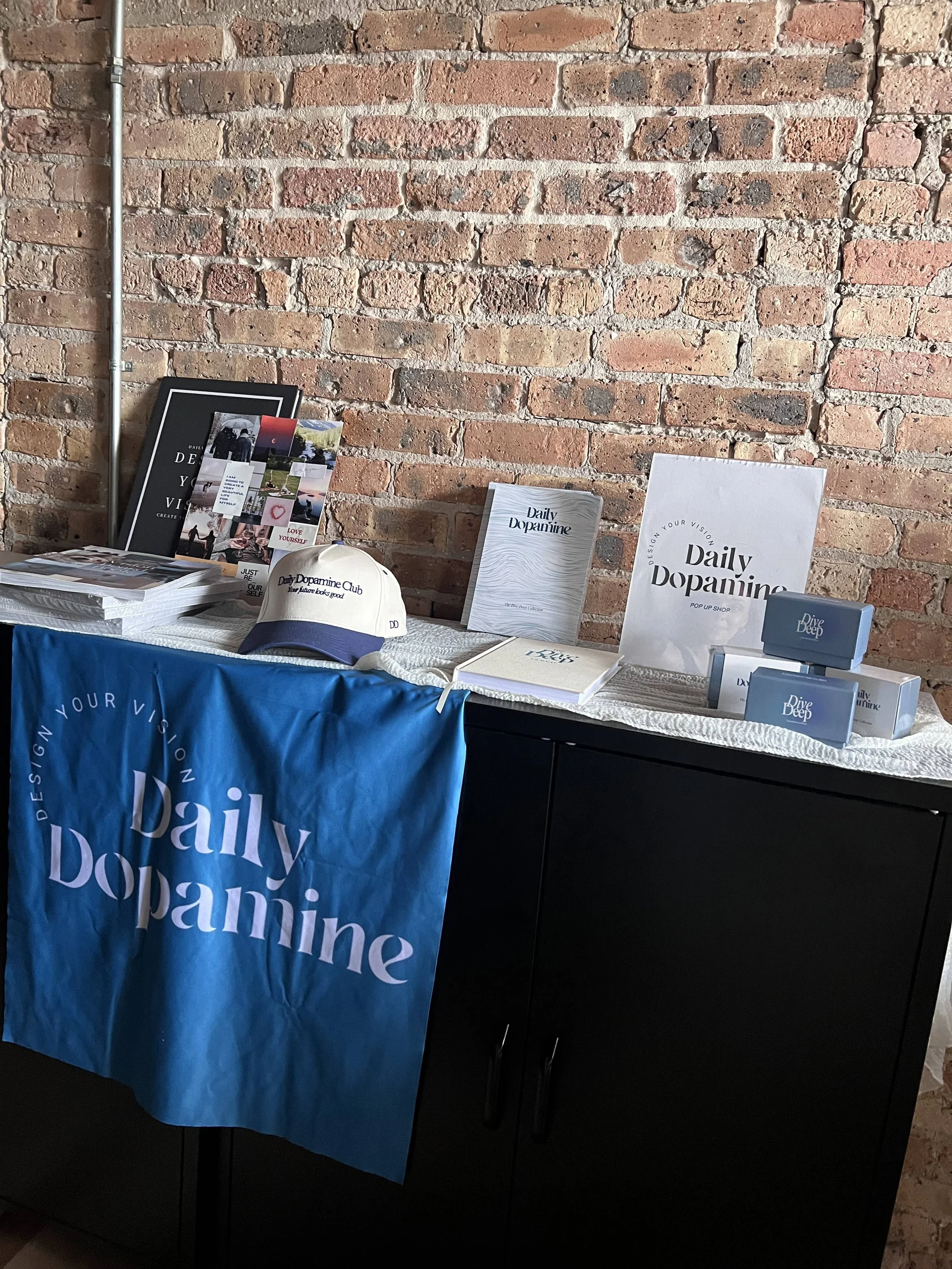 Display table with promotional materials, books, and merchandise for Daily Dopamine against an exposed brick wall. Items include a large poster, brochures, a cap, and boxes labeled 'Dive Deep' on the table, which is covered with a blue tablecloth wit