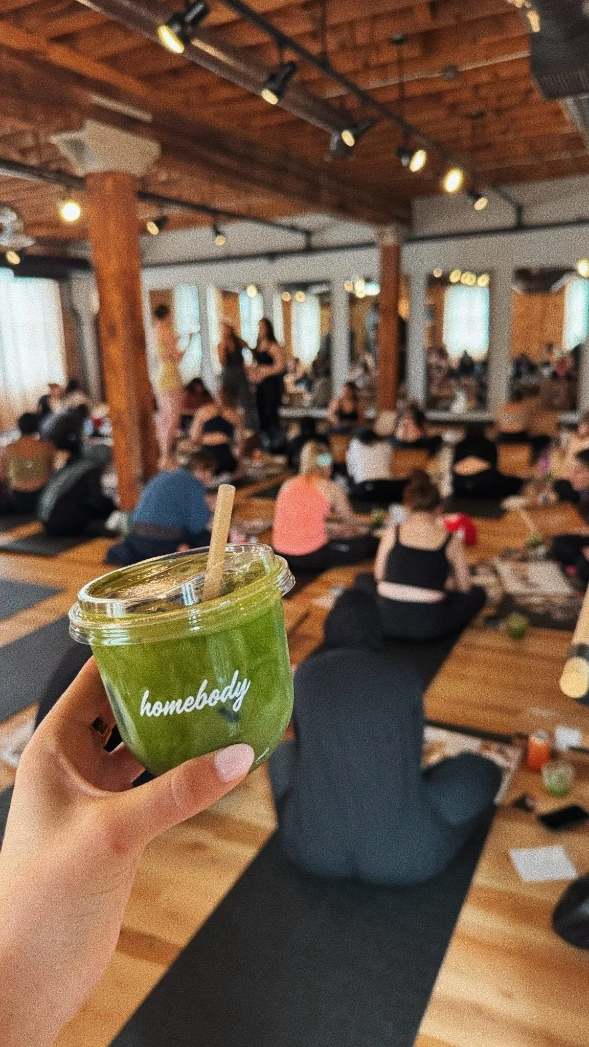 A person holding a green smoothie with a straw in a cafe or restaurant with people sitting on the floor and engaging in a group activity, with wooden beams and large windows in the background.