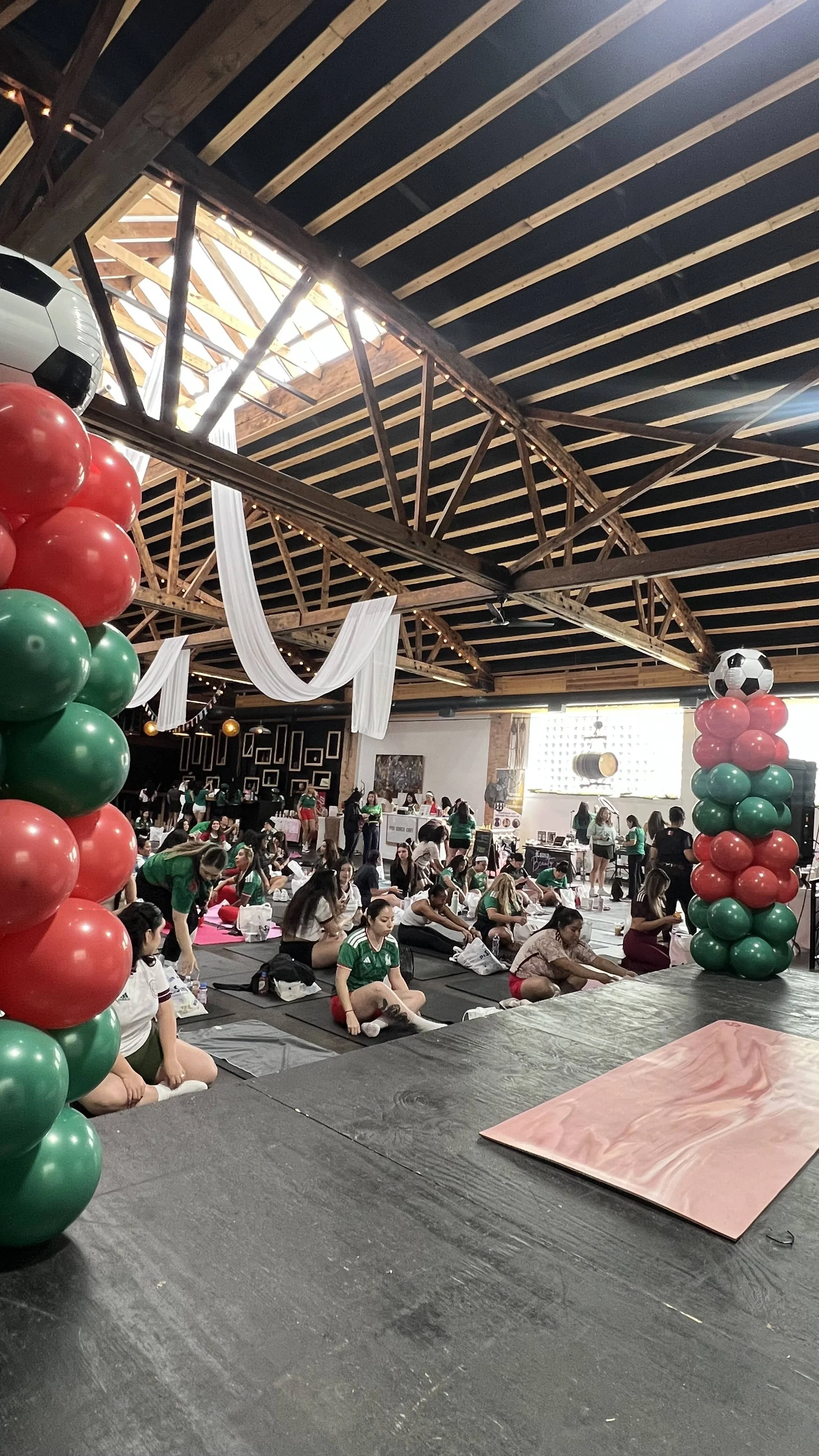 Indoor event with many women participating in a group yoga session, decorated with balloon columns with soccer ball toppers, some women wearing green shirts, wooden ceiling with skylights, white drapes, and a decorated stage area.