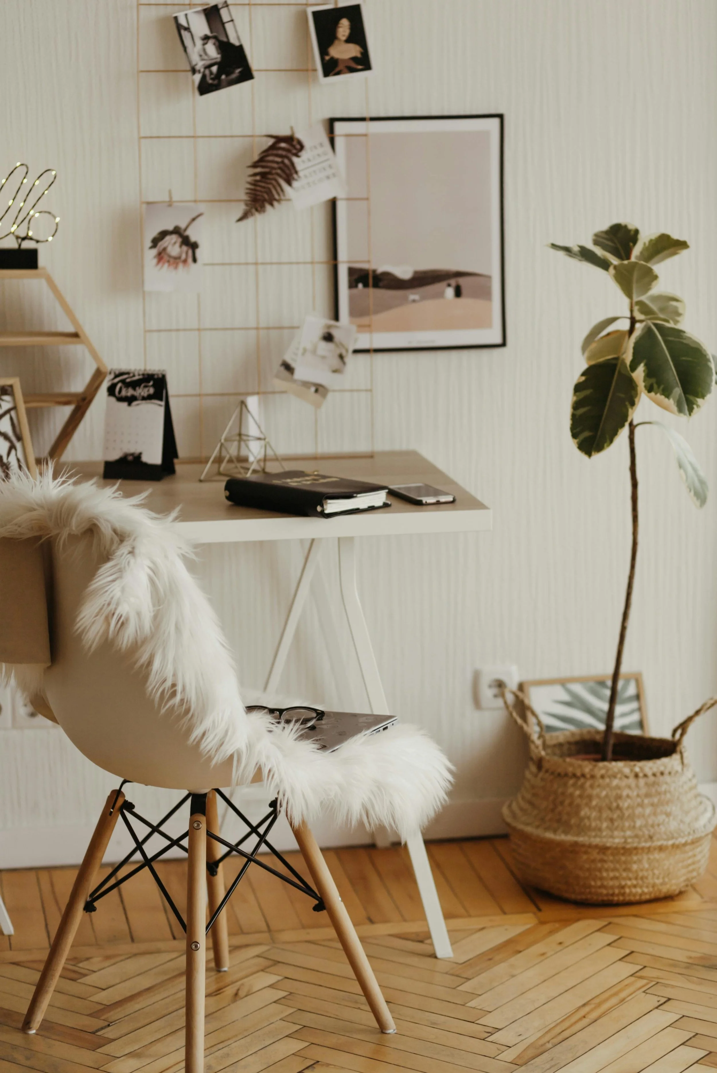 A home office setup with a wooden desk, a photo display on a wire frame, a framed landscape photograph on the wall, a small plant in a woven basket, and a white chair with a fluffy fur throw, all on a wooden parquet floor.
