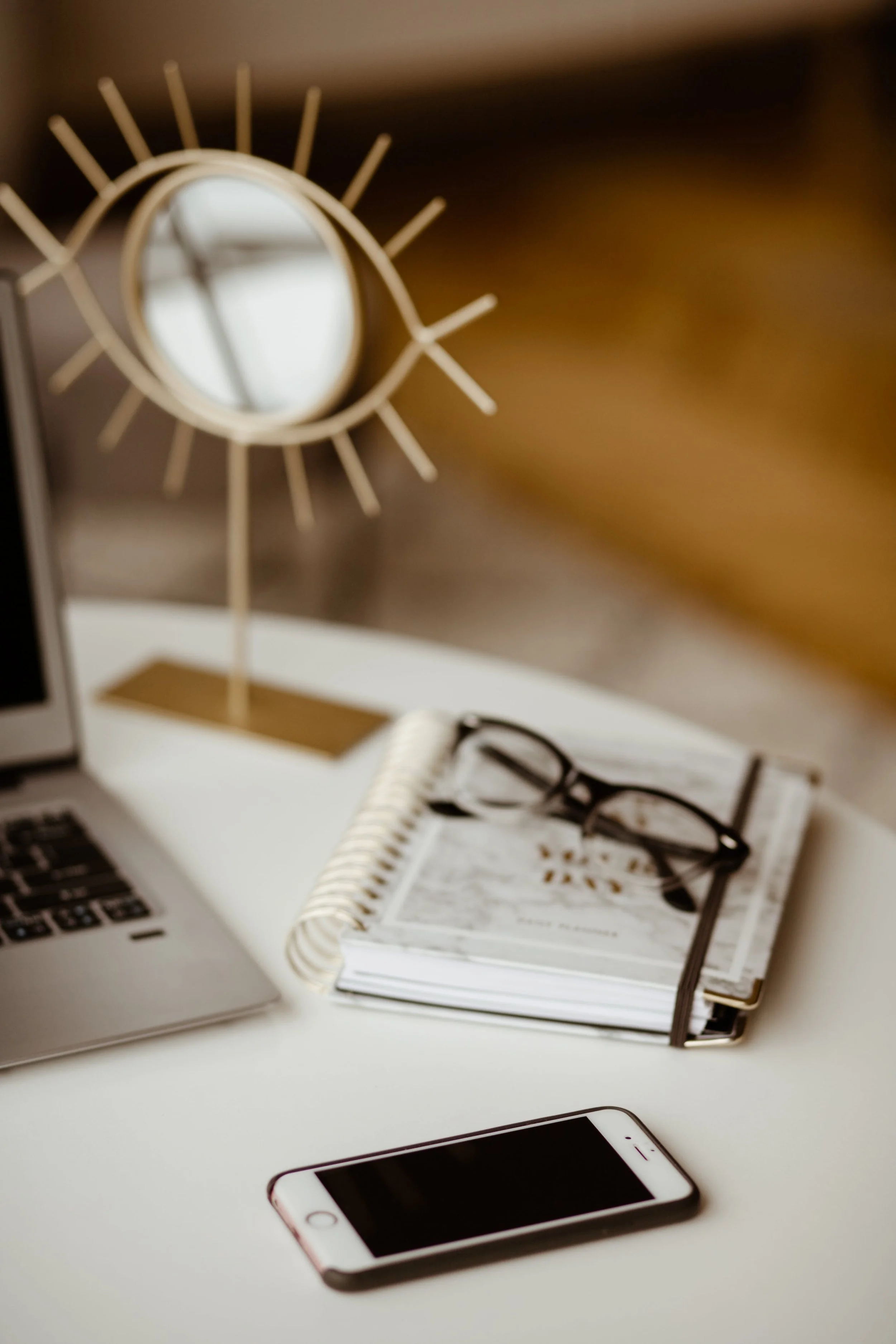 Desk with a white notebook, black eyeglasses, a smartphone, a laptop, and a decorative mirror