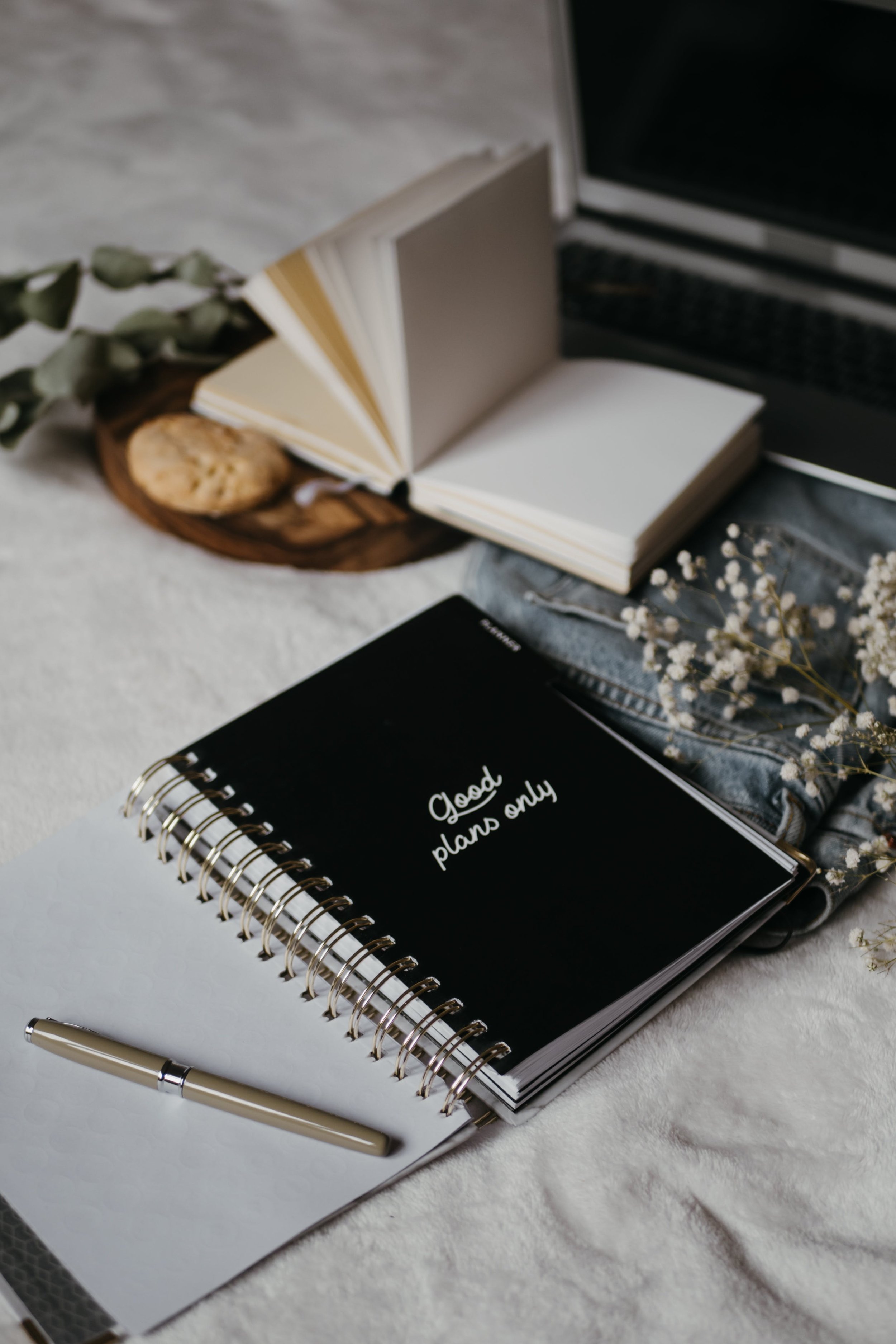 Open spiral-bound notebook with blank pages, silver pen, and black planner with white text that says 'Good plans only,' on a white textured surface, with dried flowers and a laptop in the background.