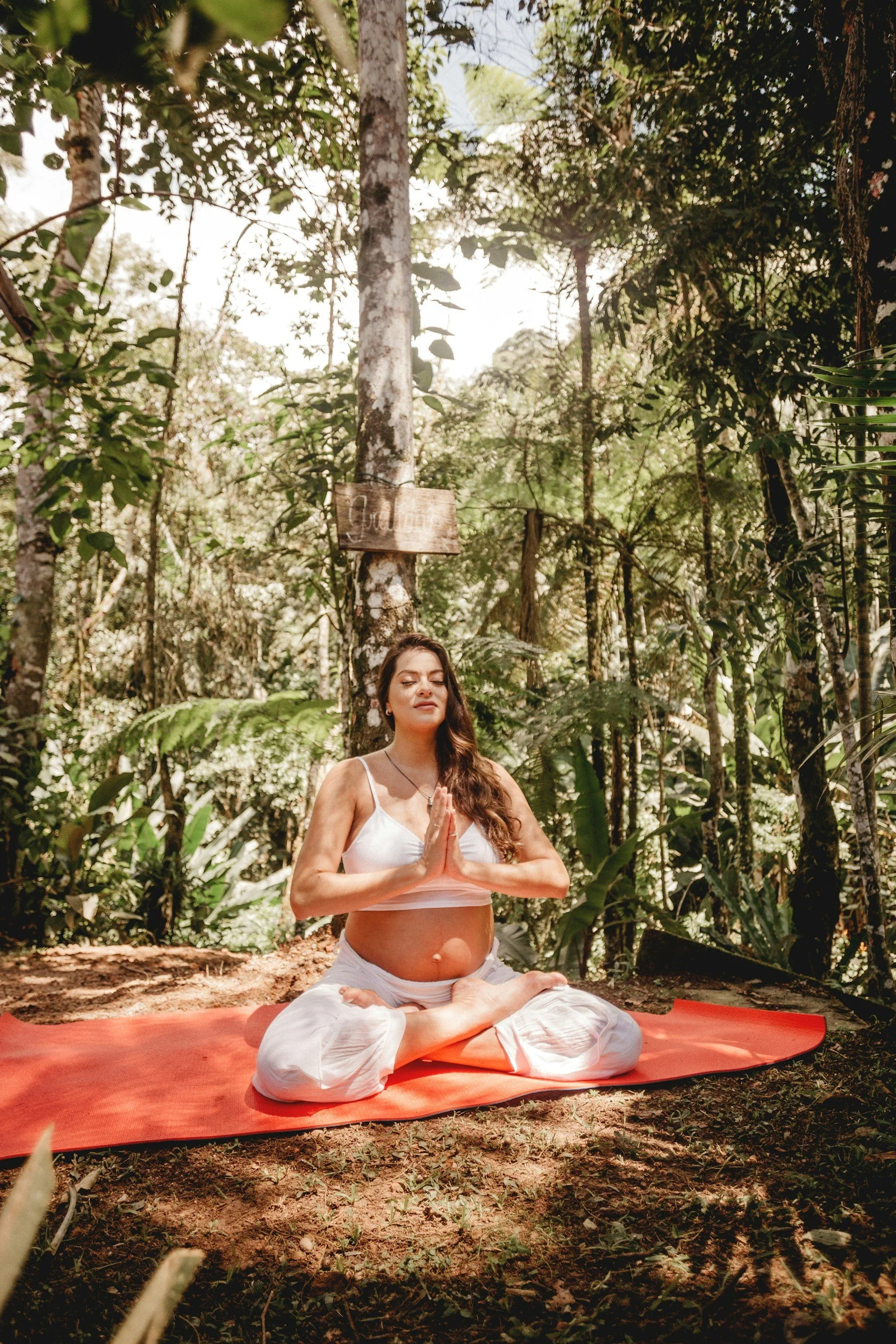 Femme enceinte faisant la méditation en position du lotus, pratiquant le yoga ou la méditation dans une forêt verdoyante, assise sur un tapis orange.