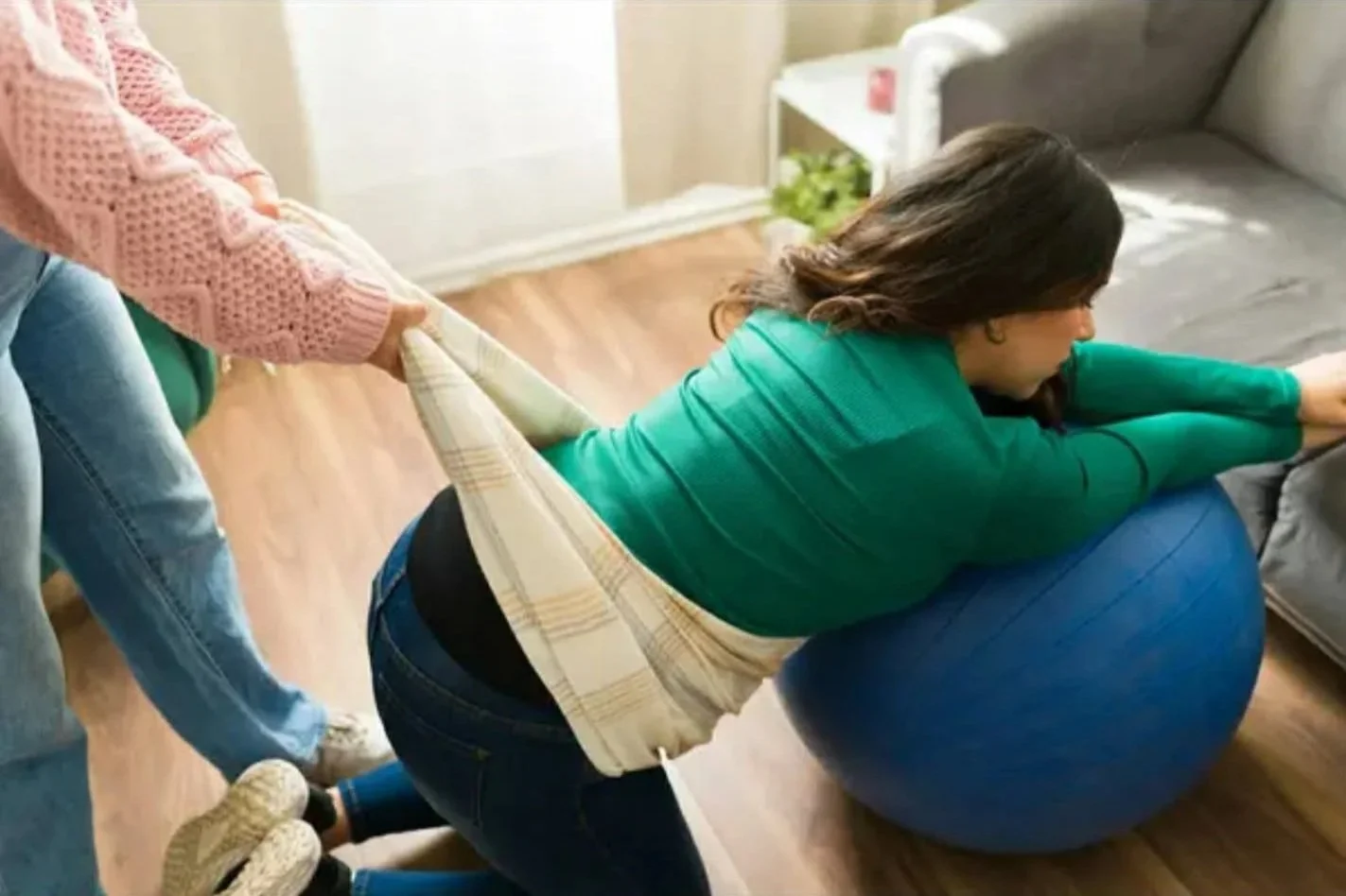 Une femme en position de départ pour faire des abdos, une autre personne la retient par sa taille dans un salon avec un canapé et un sol en parquet.