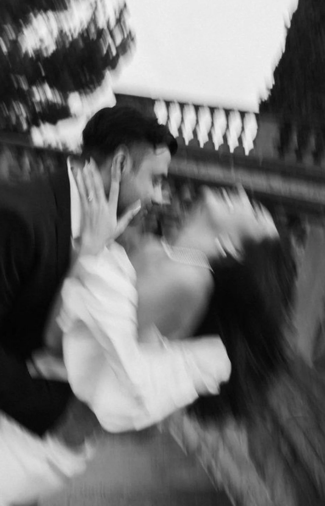 A man and woman are close together at a social event, with the woman holding her hand near her face; there are other people and decorations in the background.