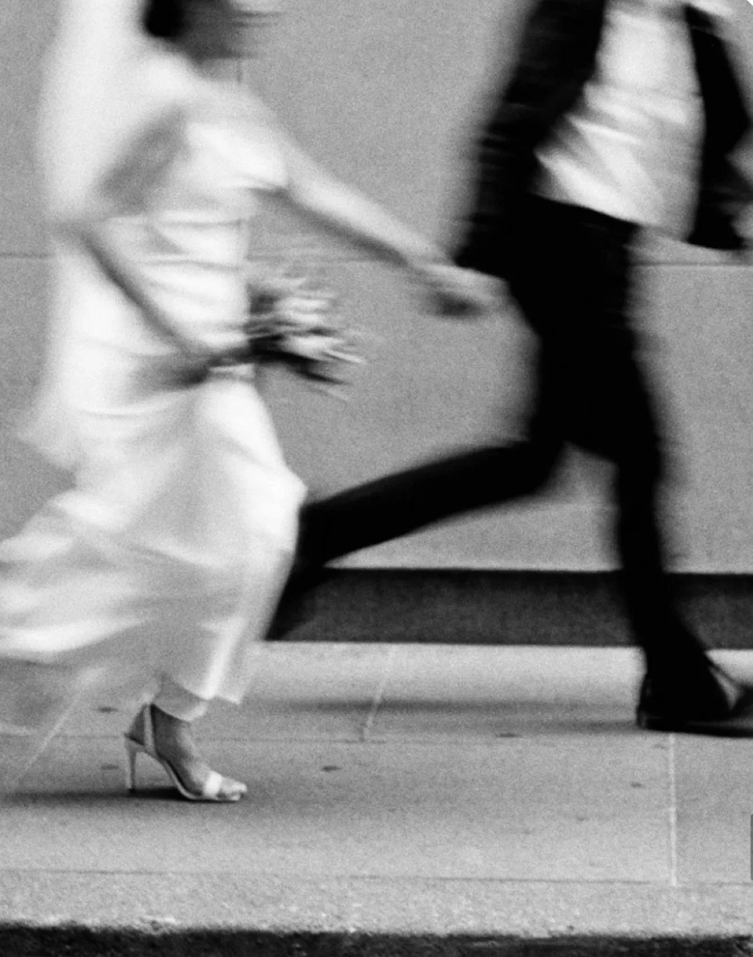Blurry black and white photo of a woman in a long dress and high heels holding hands and walking with a man in jeans and a leather jacket.