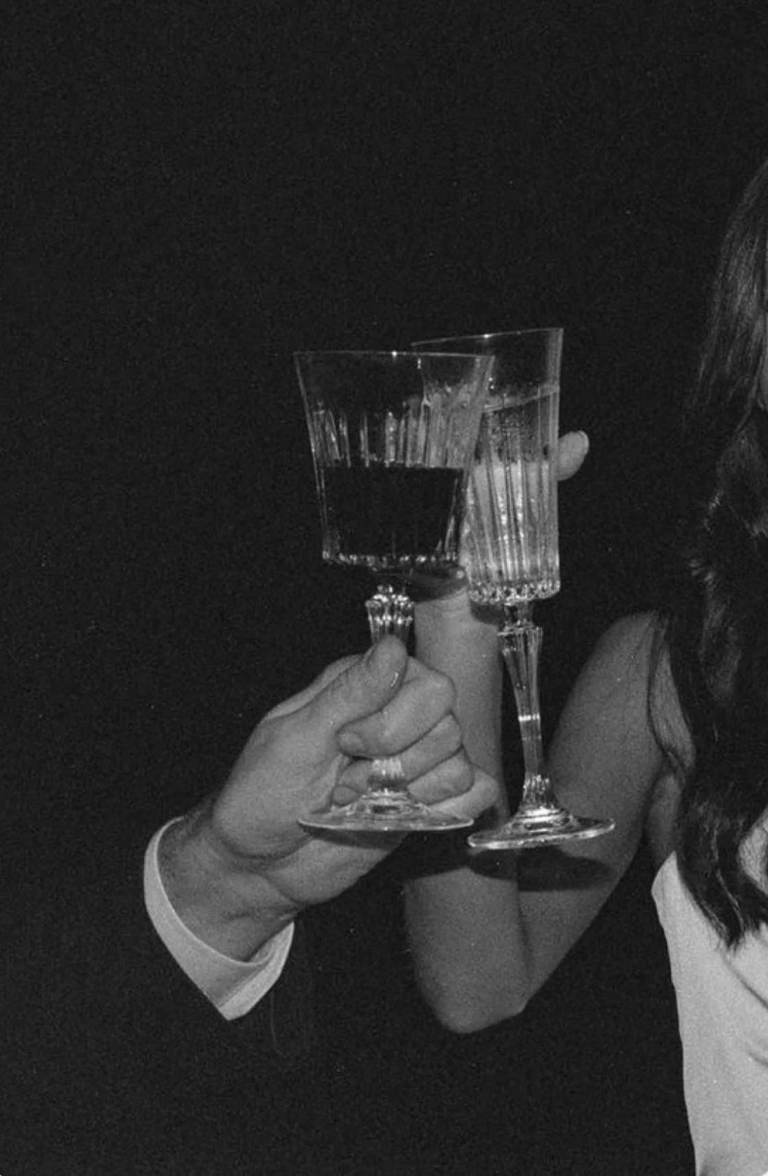 A person holding two cocktail glasses, one with a dark drink and the other with a clear drink, in a black-and-white photo.