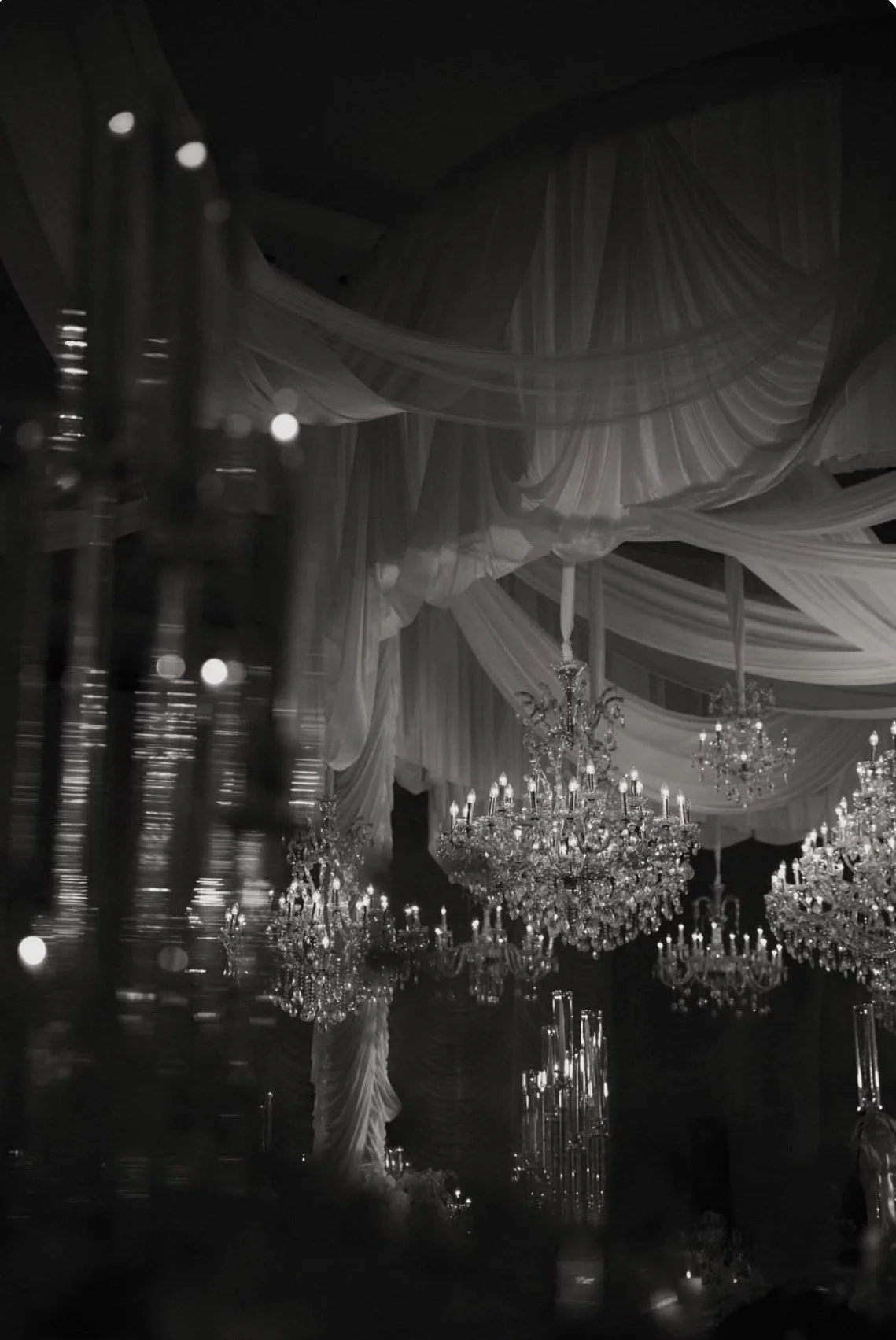 Elegant event hall decorated with hanging drapes, multiple crystal chandeliers, and soft lighting.