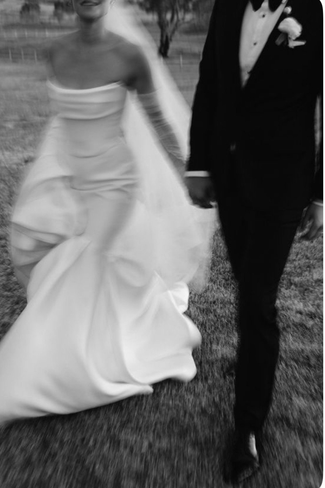 Blurred black-and-white photo of a bride and groom holding hands outdoors, the bride in a strapless gown and the groom in a tuxedo.