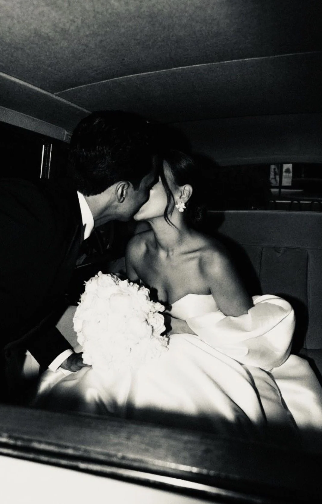 A black and white photo of a bride and groom kissing inside a vehicle. The bride is wearing a strapless wedding dress and holding a bouquet, while the groom is dressed in a tuxedo.