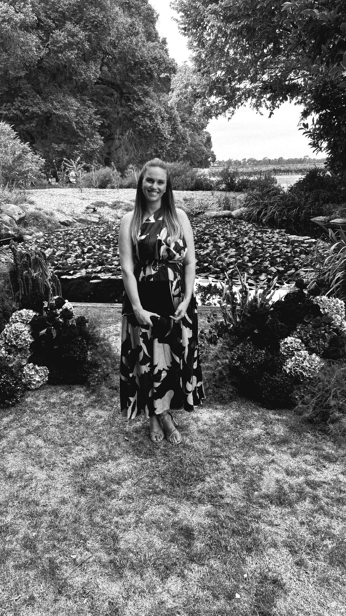 A woman in a long floral dress standing in a garden with water lilies, bushes, trees, and a pond in the background, smiling at the camera.
