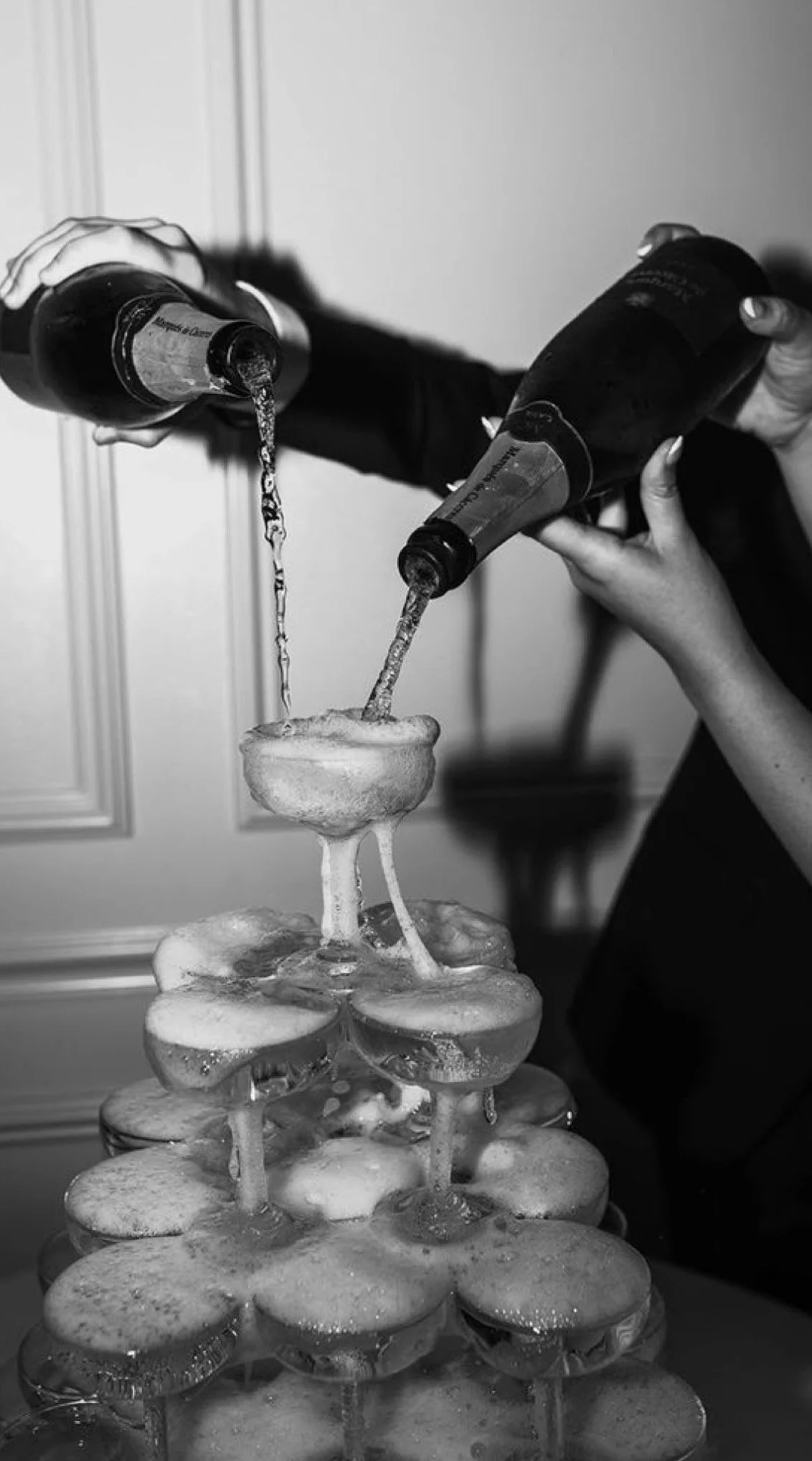 Two people are pouring liquid onto a stack of champagne glasses arranged like a fountain.