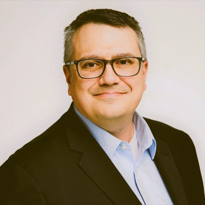 A professional man with short gray hair, glasses, wearing a light blue shirt and black blazer, smiling against a plain white background.