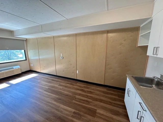 Empty room with wooden flooring, a window with sunlight, a wall with beige wood paneling, and a kitchenette with white cabinets and a sink.