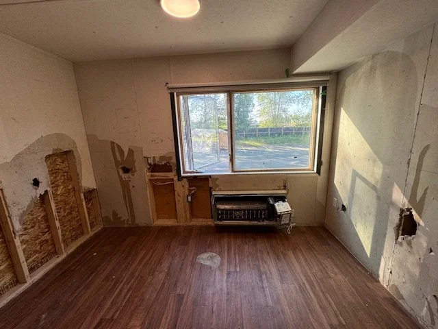 Empty room with damaged interior walls, exposed wall studs and insulation, a large window with a view of trees outside, and hardwood flooring.