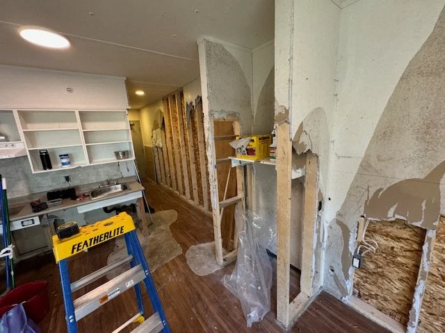 Interior view of a room under renovation with exposed wall studs, partially removed drywall, a step ladder, and construction tools.
