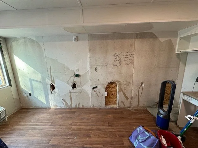 Living room with damaged wall showing holes and torn drywall, hardwood floor, window on left, baseboards, fireplace vent, and miscellaneous items including a Dyson fan and bags.