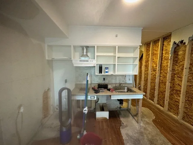 Small kitchen area under renovation with exposed wall studs on the right, a white cabinet with open shelves and a range hood above a small countertop and sink. A vacuum cleaner and mop are in the foreground.