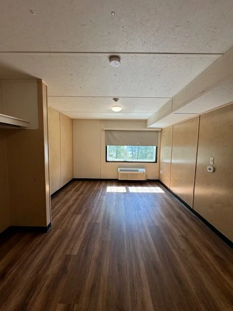 Empty room with wood flooring, beige walls, a window with a cover, and an air conditioning unit underneath the window.