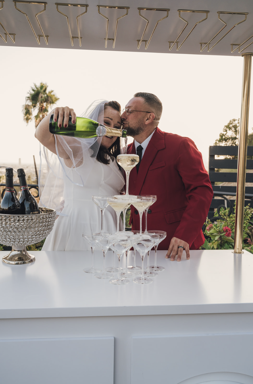 A newlywed couple sharing a champagne toast at their wedding reception, with the groom in a red suit and the bride in a white dress, pouring champagne into a stack of glasses.