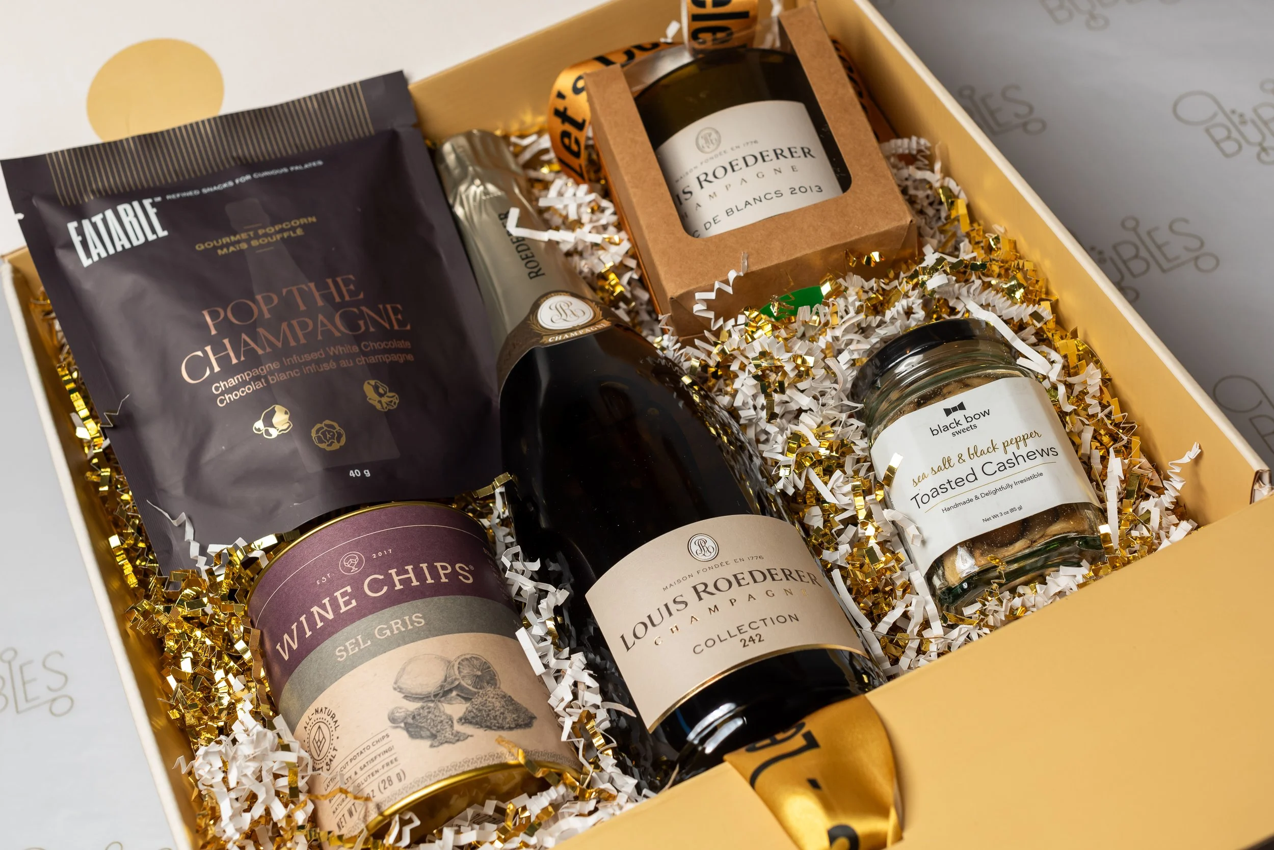 A gift box filled with gourmet snacks, including a bag of popcorn, a bottle of Louis Roederer champagne, a jar of toasted cashews, and a small bottle of champagne, all nestled in white and gold shredded paper.
