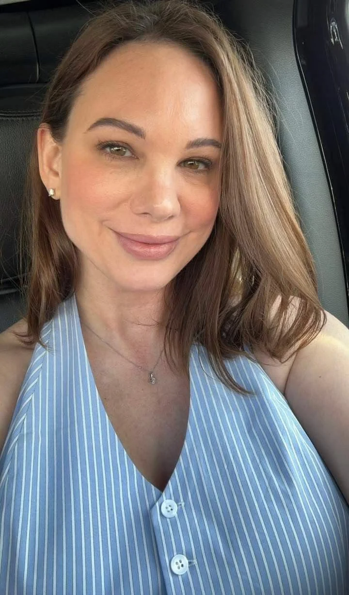 A woman with light skin and brown hair smiling, wearing a blue and white striped sleeveless top, earrings, and a necklace, sitting in a vehicle.