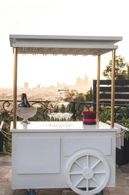 A white outdoor mobile bar cart with a canopy, set on a patio with a view of a city skyline in the background. The cart has drinks and glasses on top.