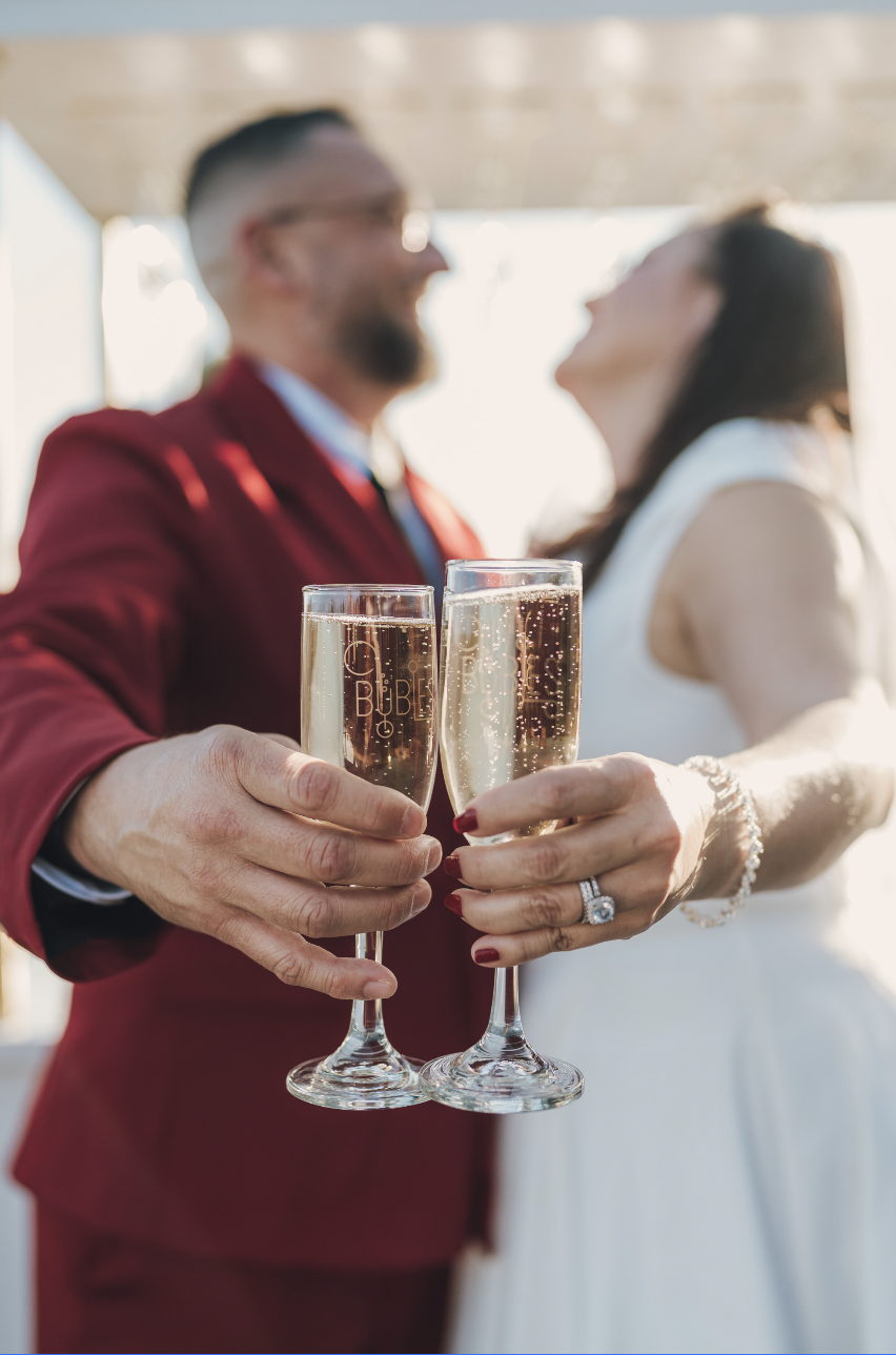 Couple at a wedding toast with champagne glasses, wearing wedding attire, with blurred bride and groom in background.