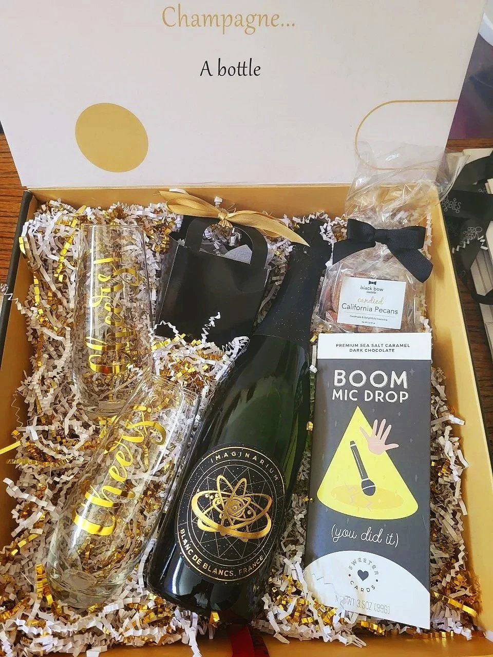 Gift box containing a bottle of sparkling wine, two champagne glasses with gold 'Cheers' lettering, a small black gift bag, a bag of candied California pecans, a box of caramel dark chocolate, and a humorous microphone-themed greeting card.