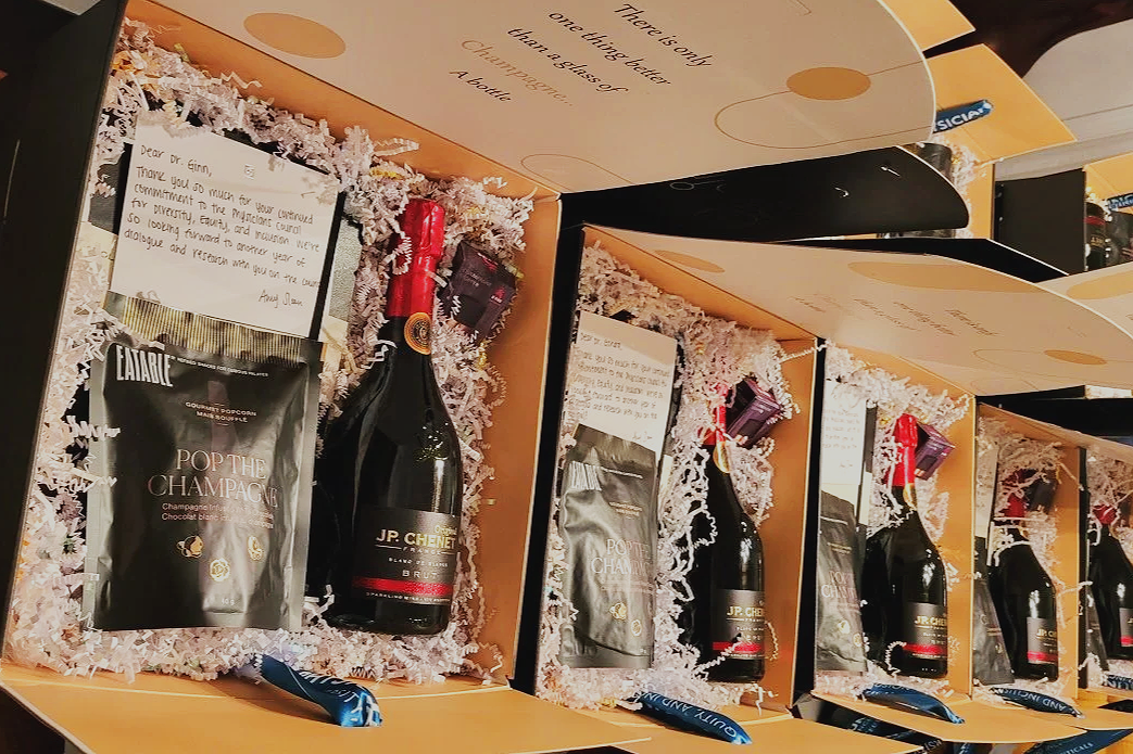 Gift boxes containing a bottle of JP Chene champagne, a bag of popcorn labeled 'Pop the Champagne,' a small chocolate bar, and a card with a thank you note.