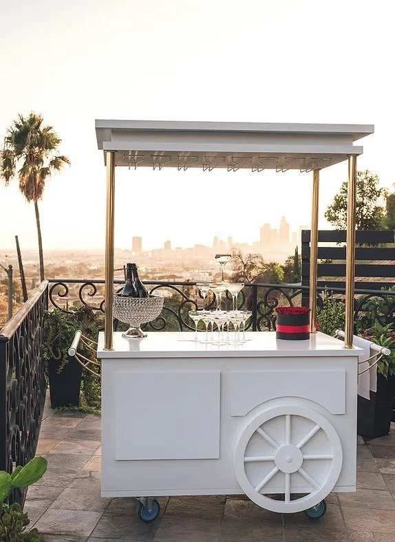 White mobile bar cart with a canopy, bottles of champagne, glasses, and a flower arrangement, set on an outdoor patio with plants, a city skyline, and a palm tree in the background.
