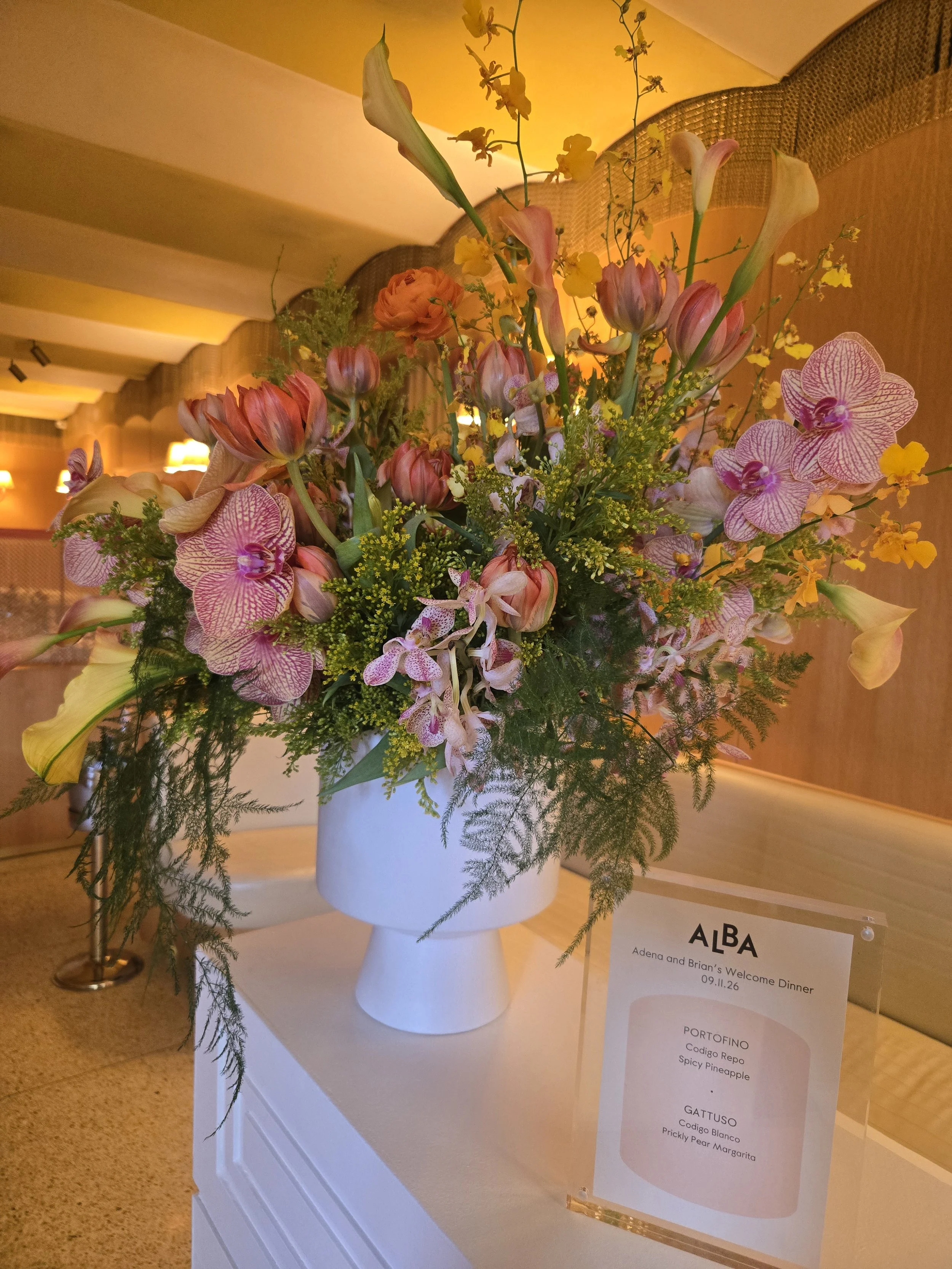 A floral arrangement in a white vase on a white stand. The arrangement includes pink and white orchids, orange ranunculus, yellow flowers, and greenery. There is a sign beside the arrangement with details about the event and the flowers used.