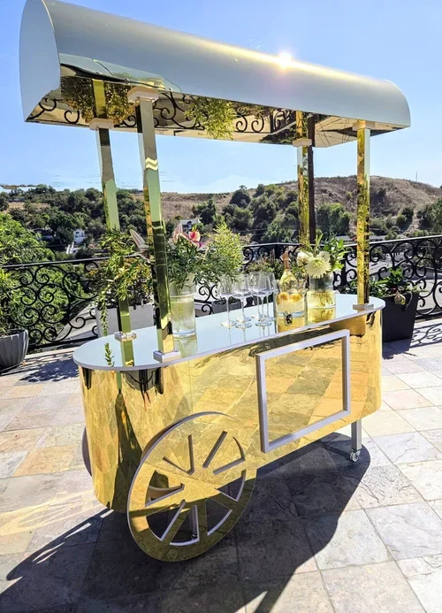 A gold mobile bar cart with a canopy on a tiled outdoor patio, decorated with flowers and drinkware, overlooking a hillside landscape.